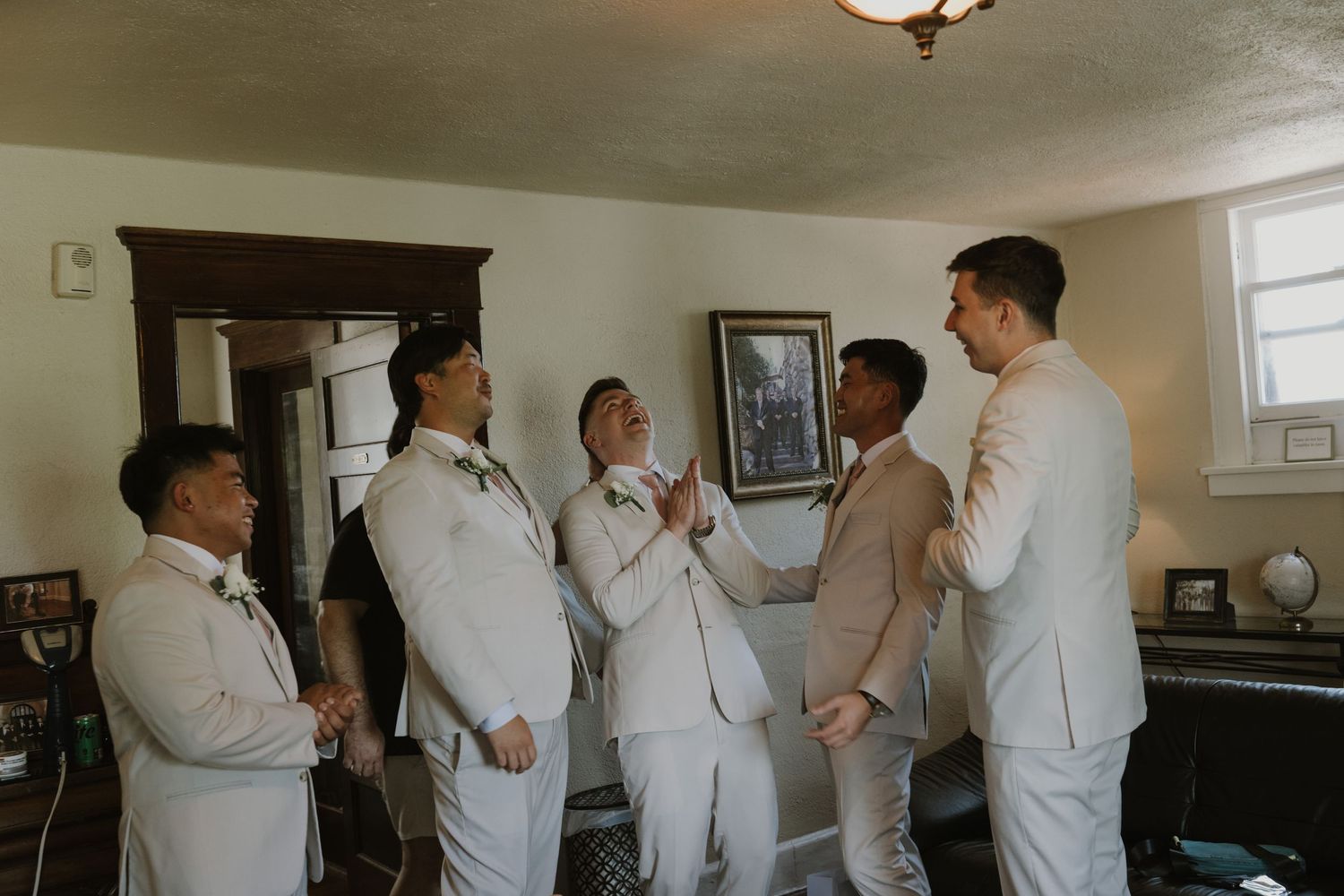 Groomsmen in white suits help each other prepare before a wedding ceremony indoors.