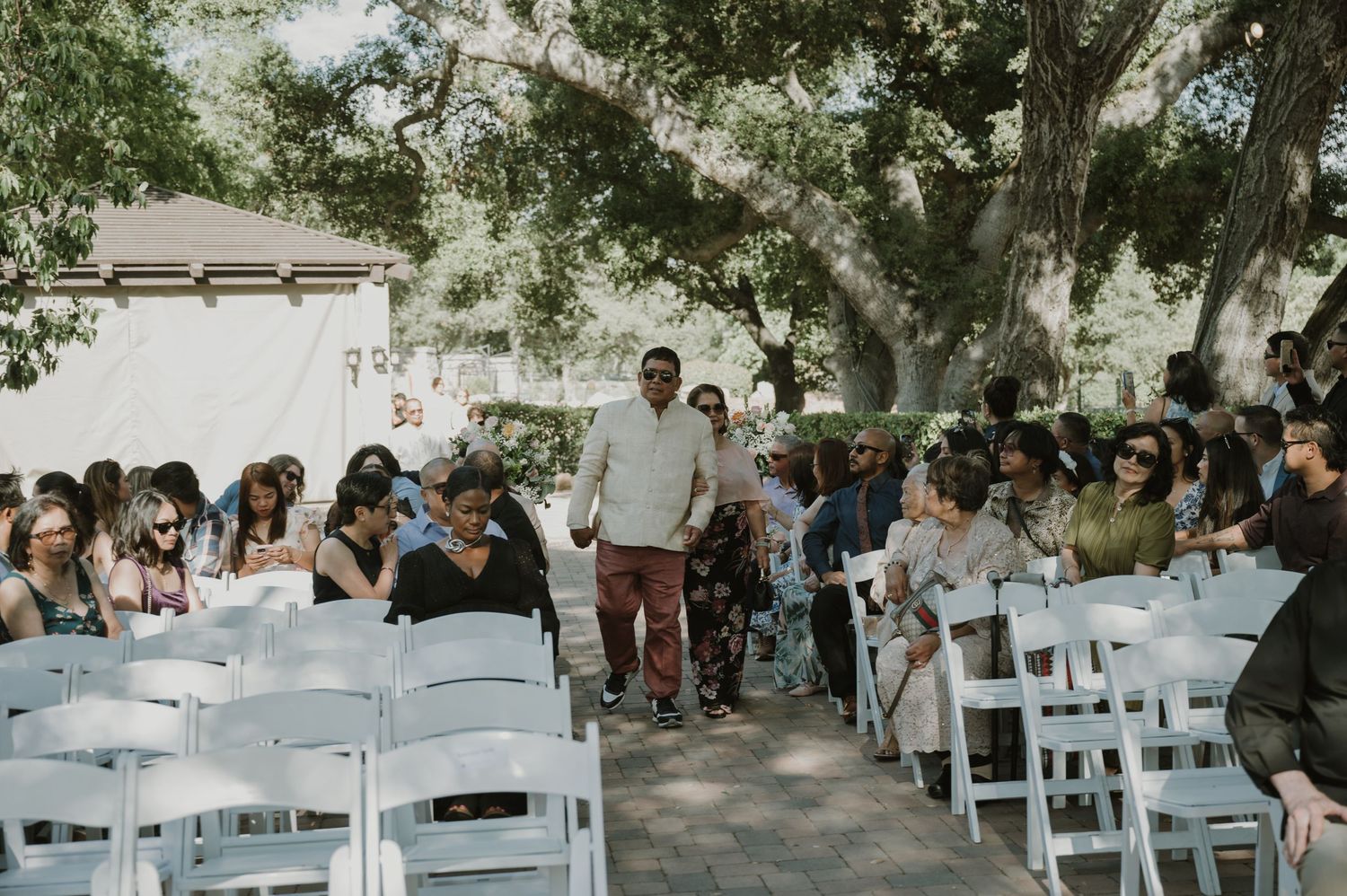 Man walks down outdoor wedding aisle lined with white chairs and seated guests under large oak trees.
