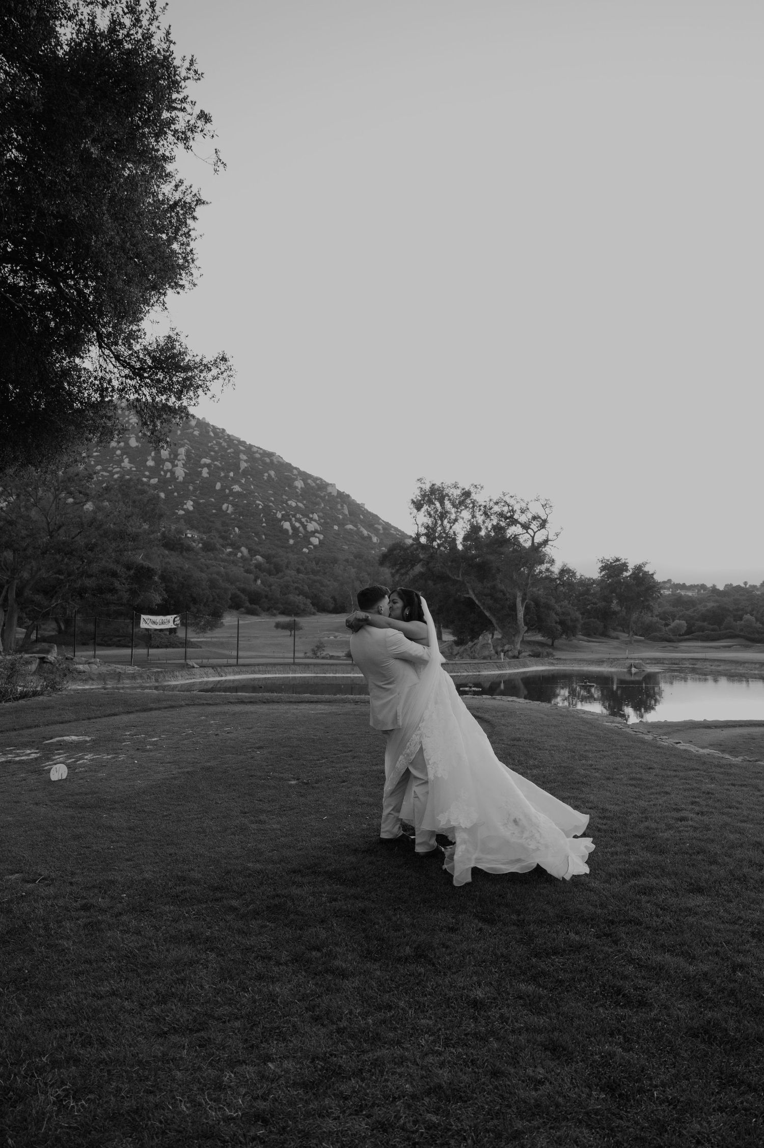 Black and white photo of groom lifting bride in flowing gown near a scenic pond.