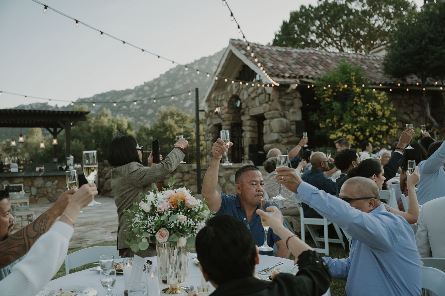 Wedding guests raise champagne glasses in a toast at an outdoor reception with string lights.