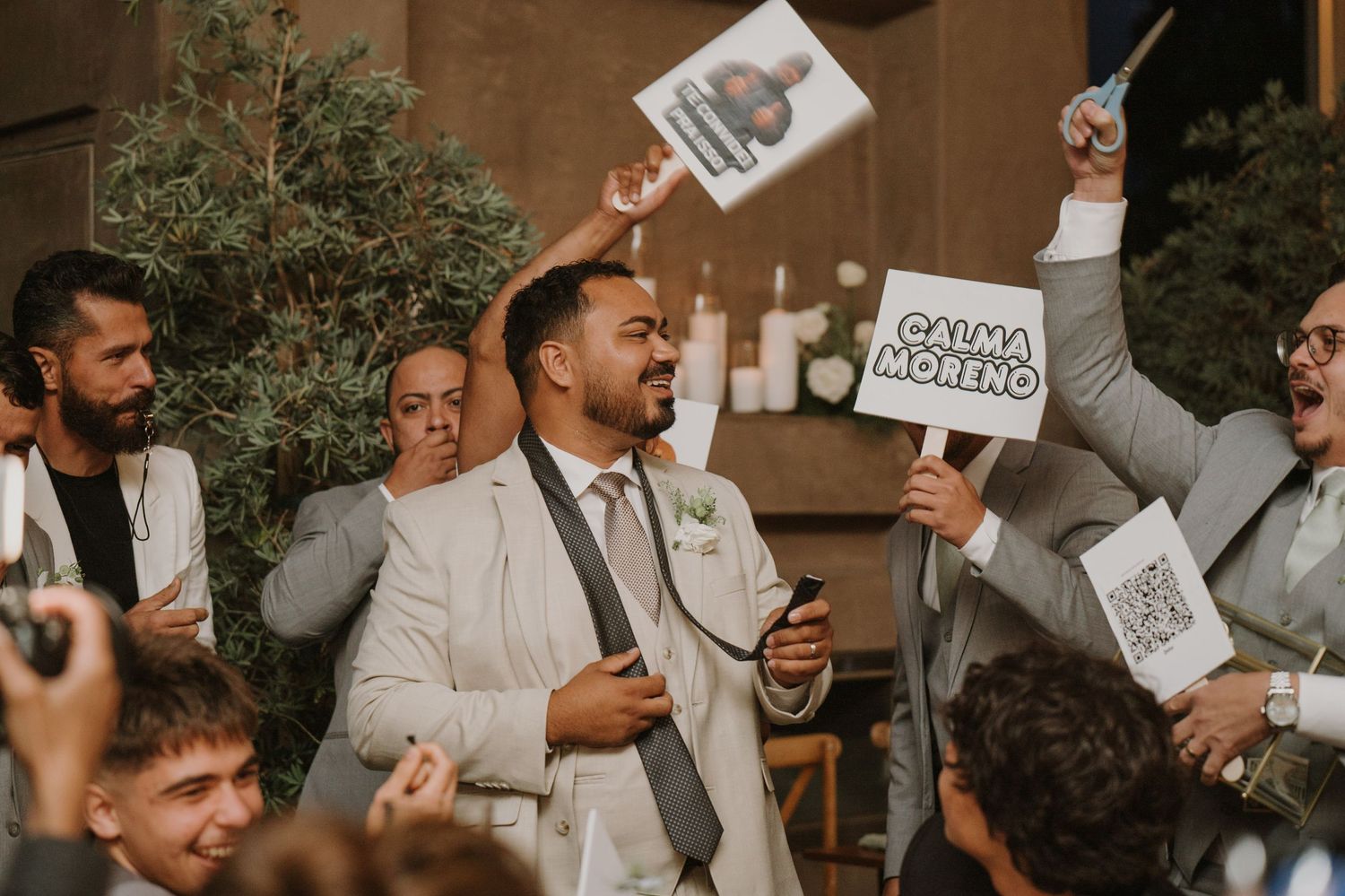 Groom in beige suit celebrating with groomsmen holding signs at lively wedding reception.