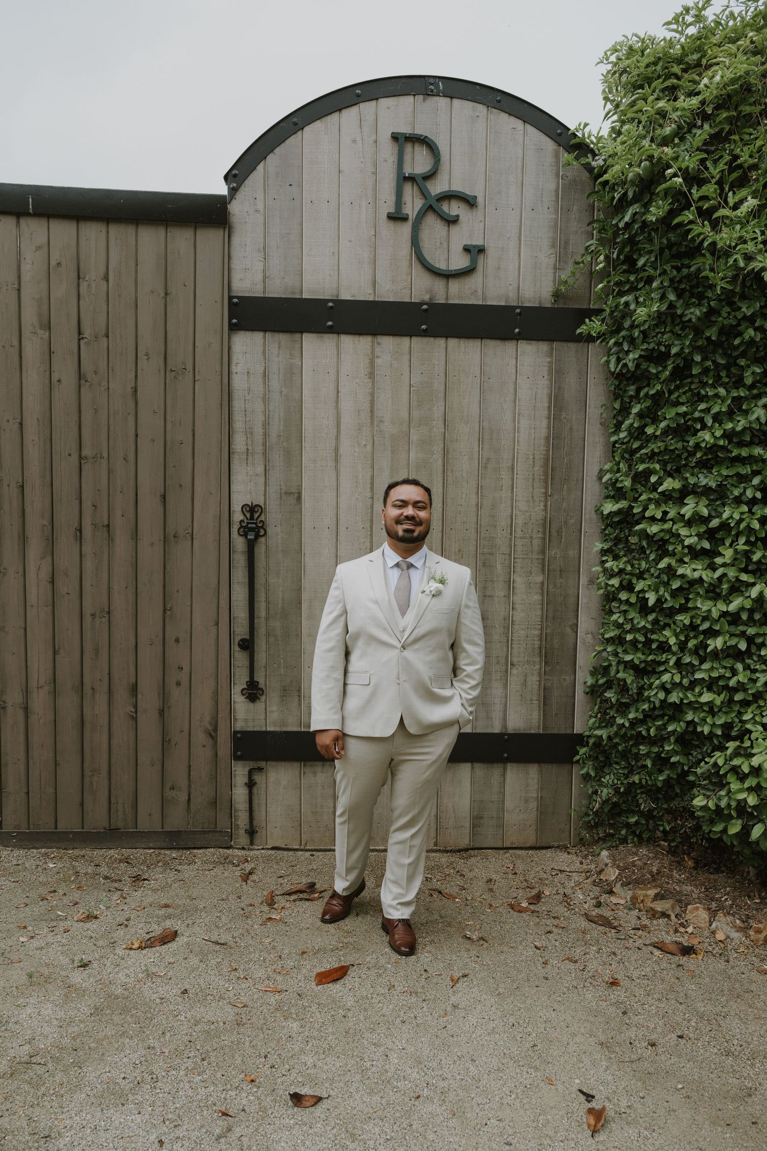 Groom in a beige suit with boutonniere stands before rustic barn doors with RC monogram at an outdoor wedding venue.