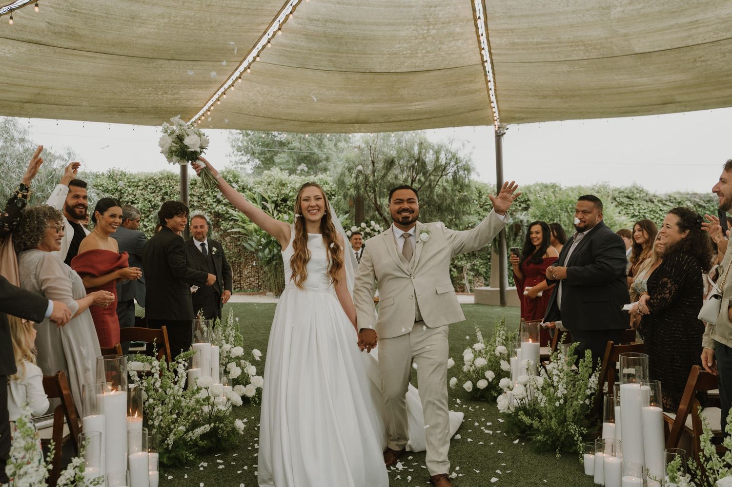 Newlyweds celebrate with raised arms as guests cheer along floral aisle under tent.