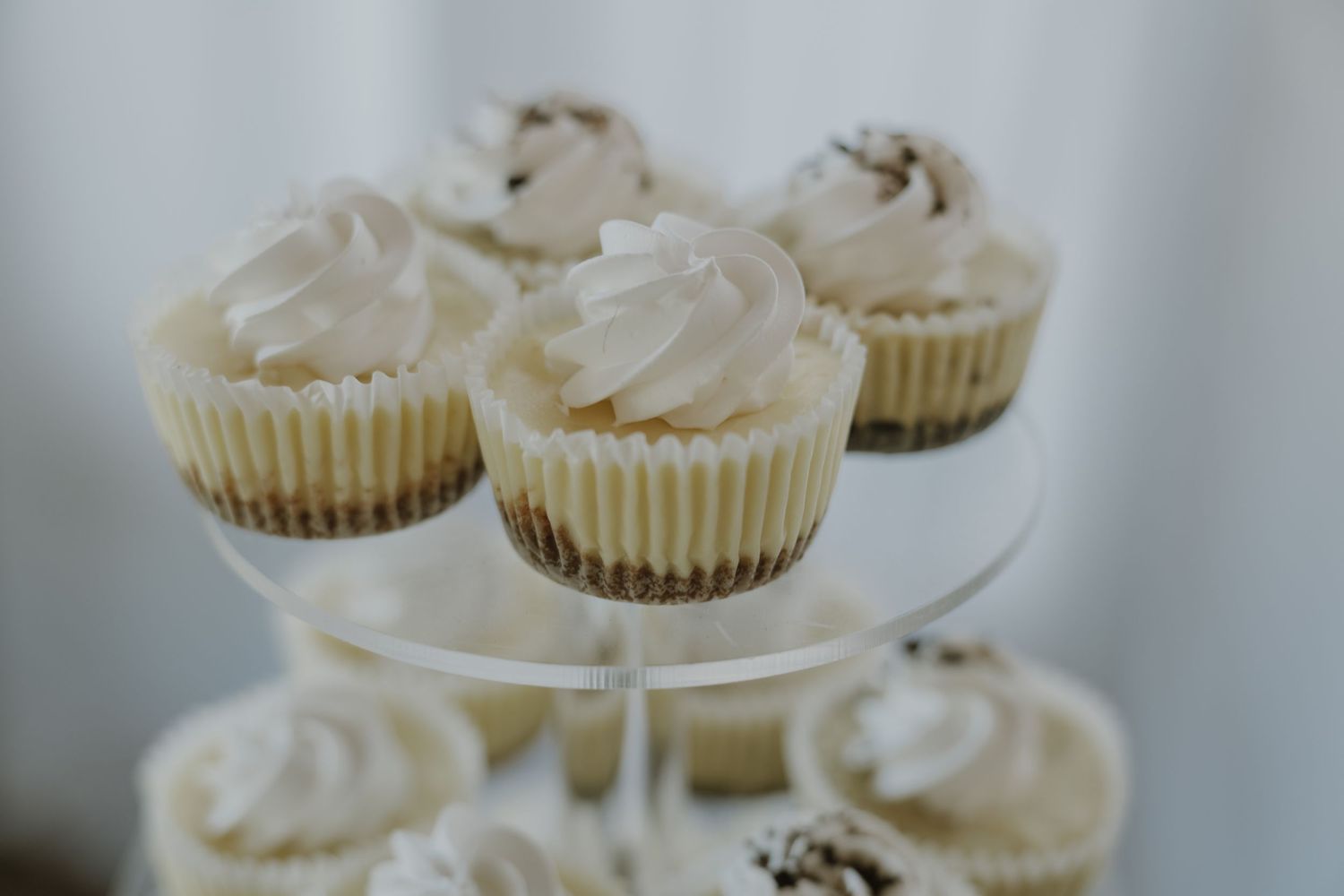 Mini cheesecakes with whipped cream topping displayed on a clear tiered dessert stand.