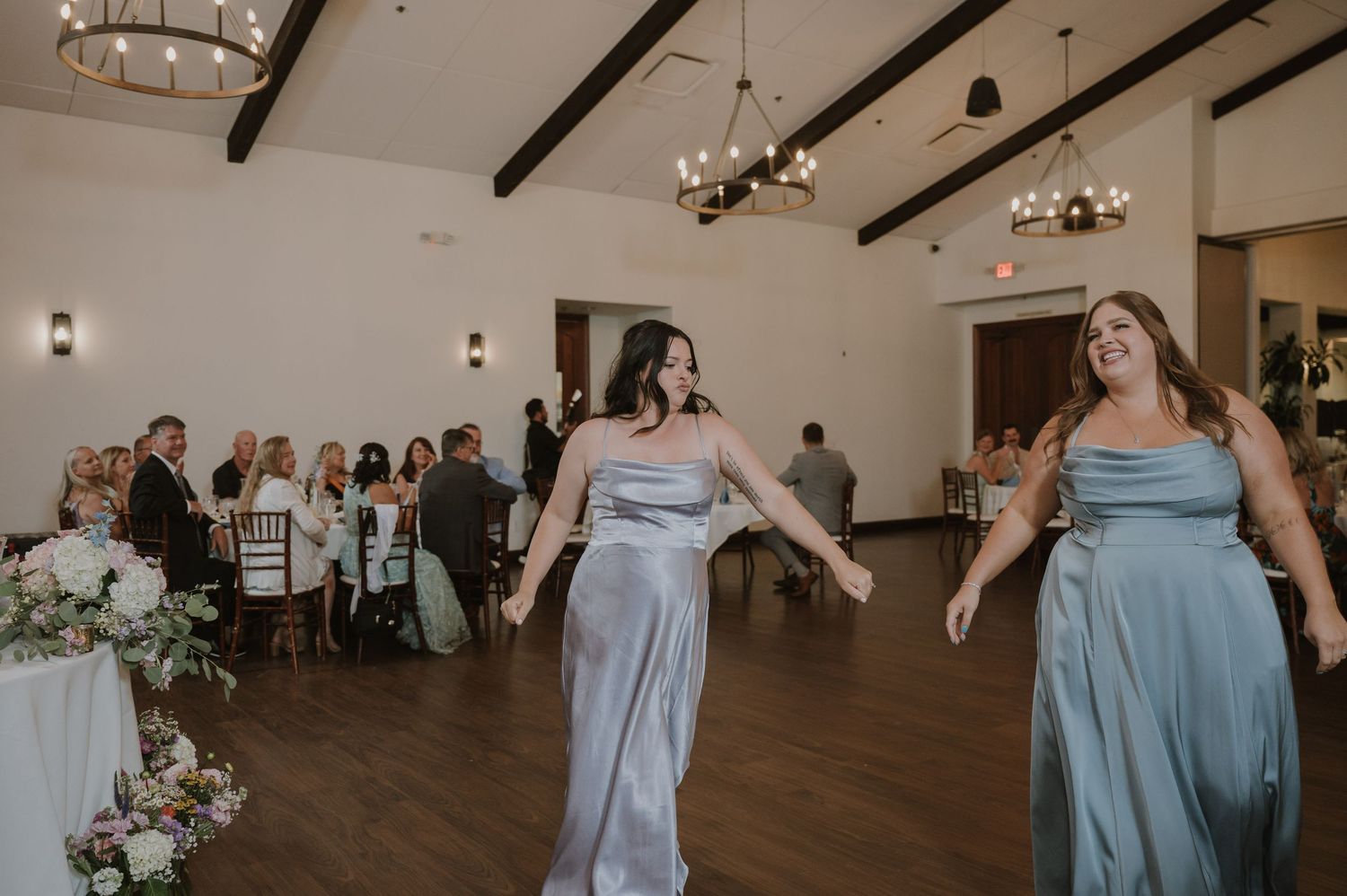Two bridesmaids in silver-blue satin dresses dancing at an elegant wedding reception venue with exposed beam ceilings.