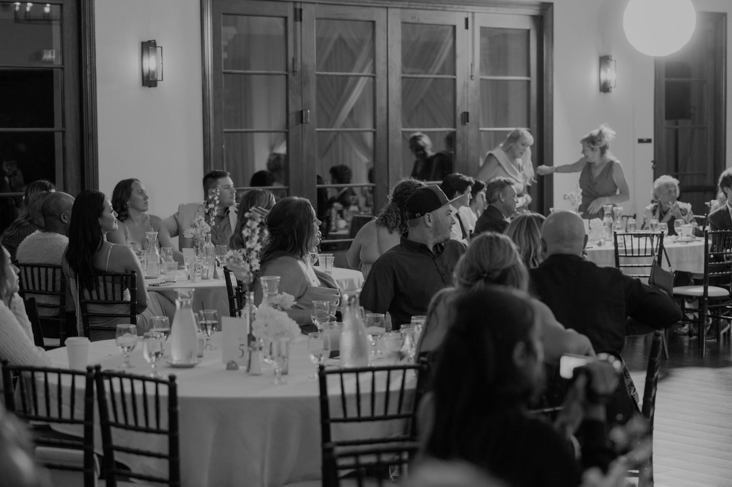 Black and white photo of wedding reception guests seated at round tables in elegant venue.