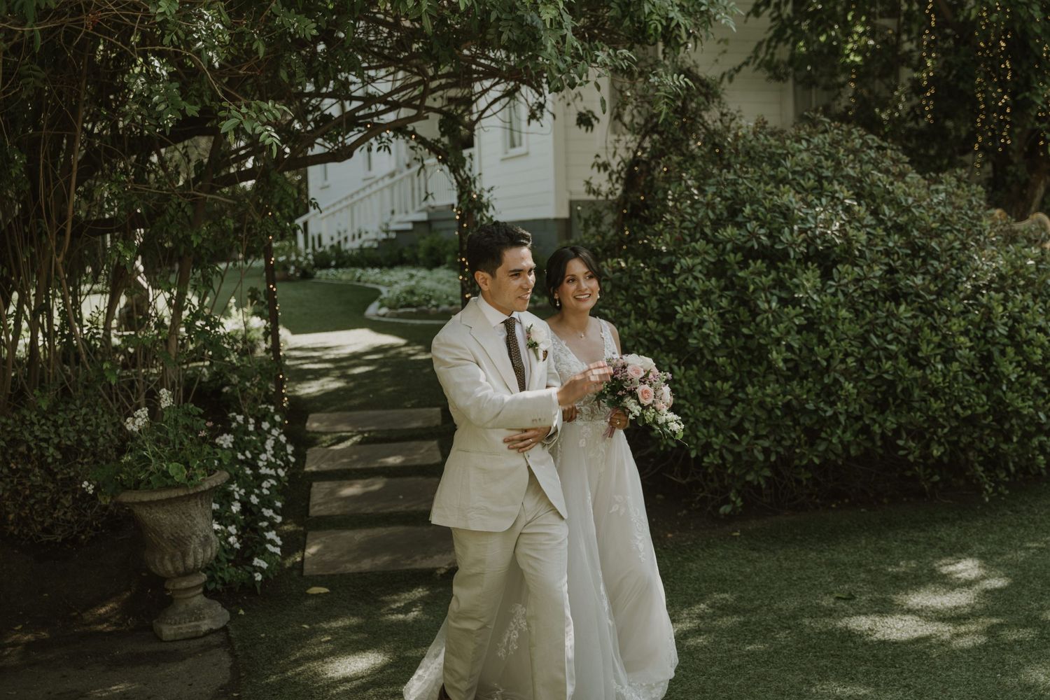 A bride and groom walk hand in hand through a lush garden, she in a white gown holding a bouquet, he in a beige suit.