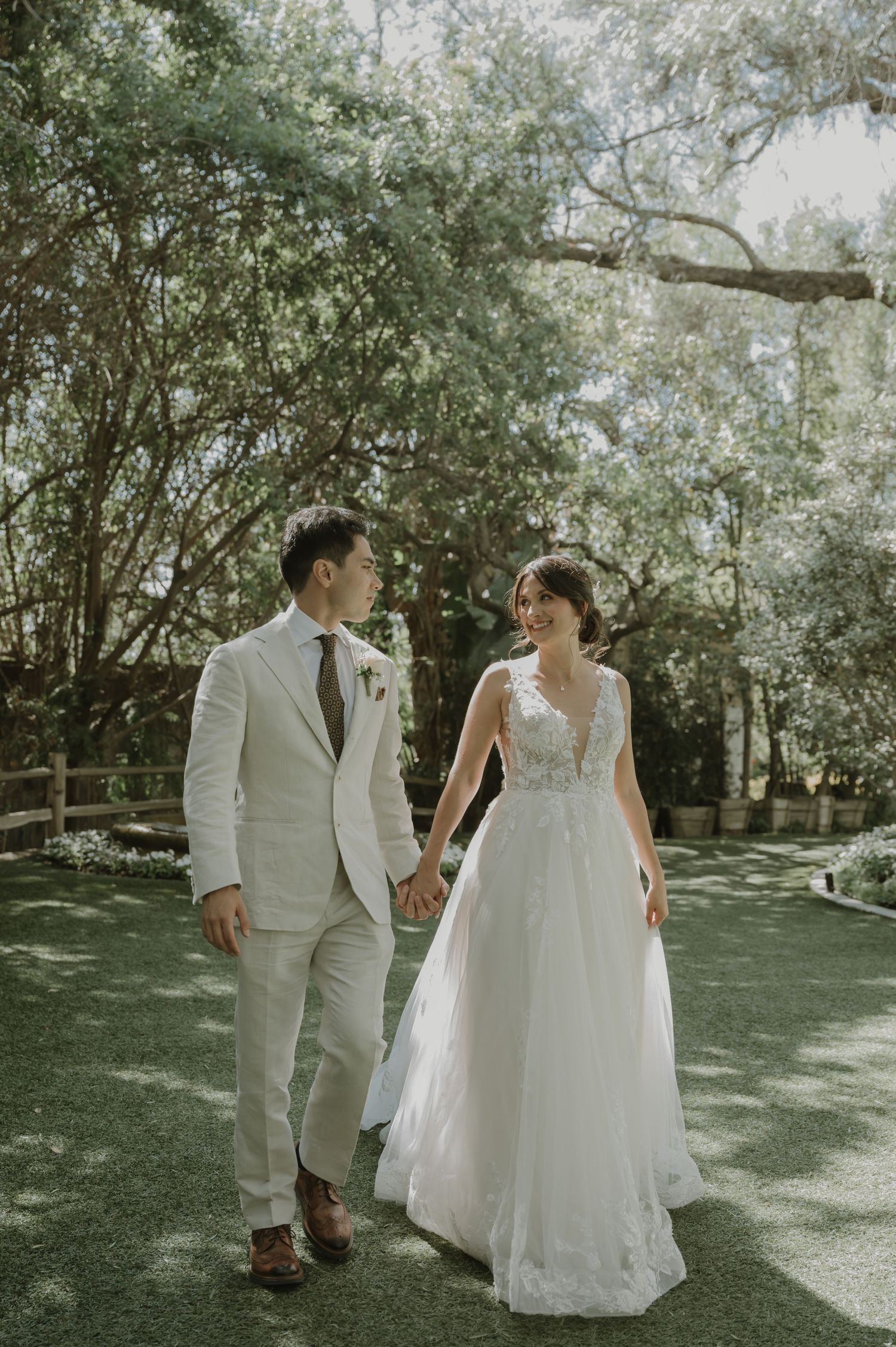 Bride and groom walk hand in hand through a sunlit garden in elegant wedding attire.