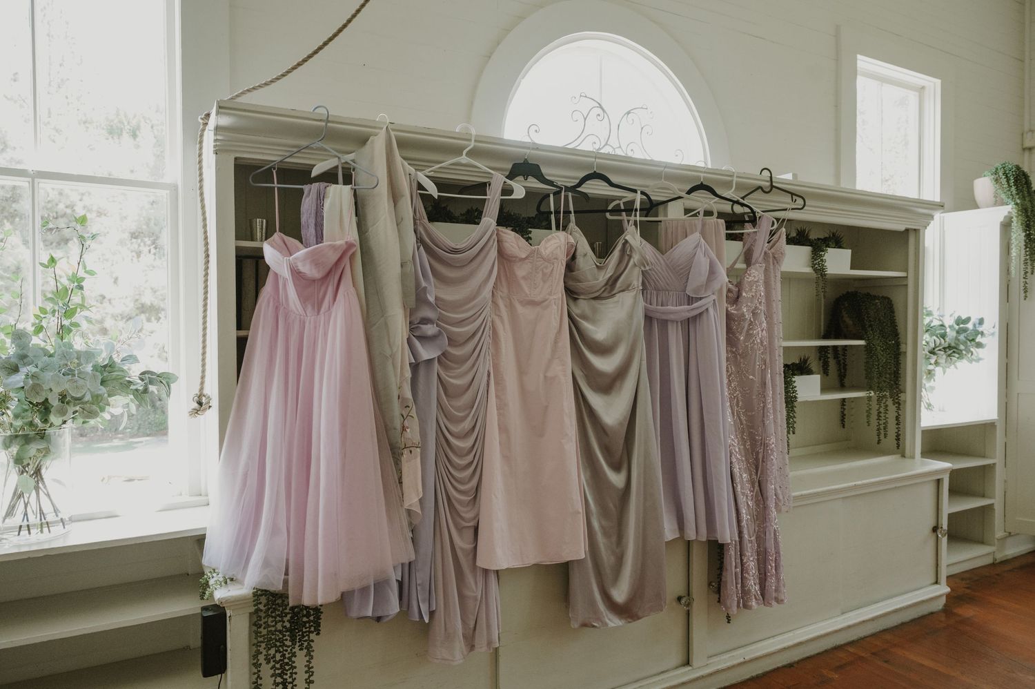 Elegant bridesmaid dresses in soft pink and lavender hues hanging on a vintage white rack in a bright bridal suite.