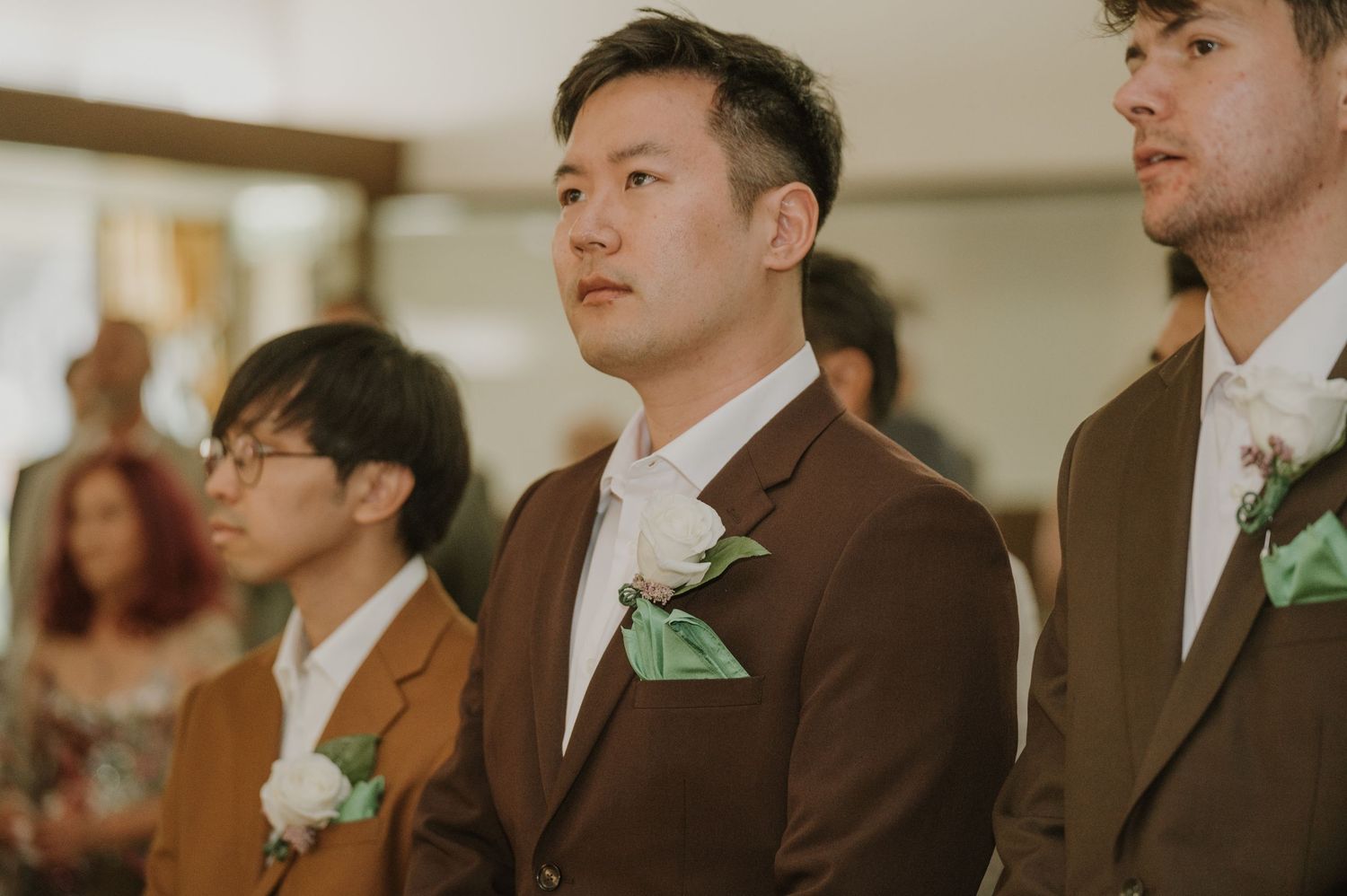 Groom in brown suit with boutonniere stands at the altar during wedding ceremony.