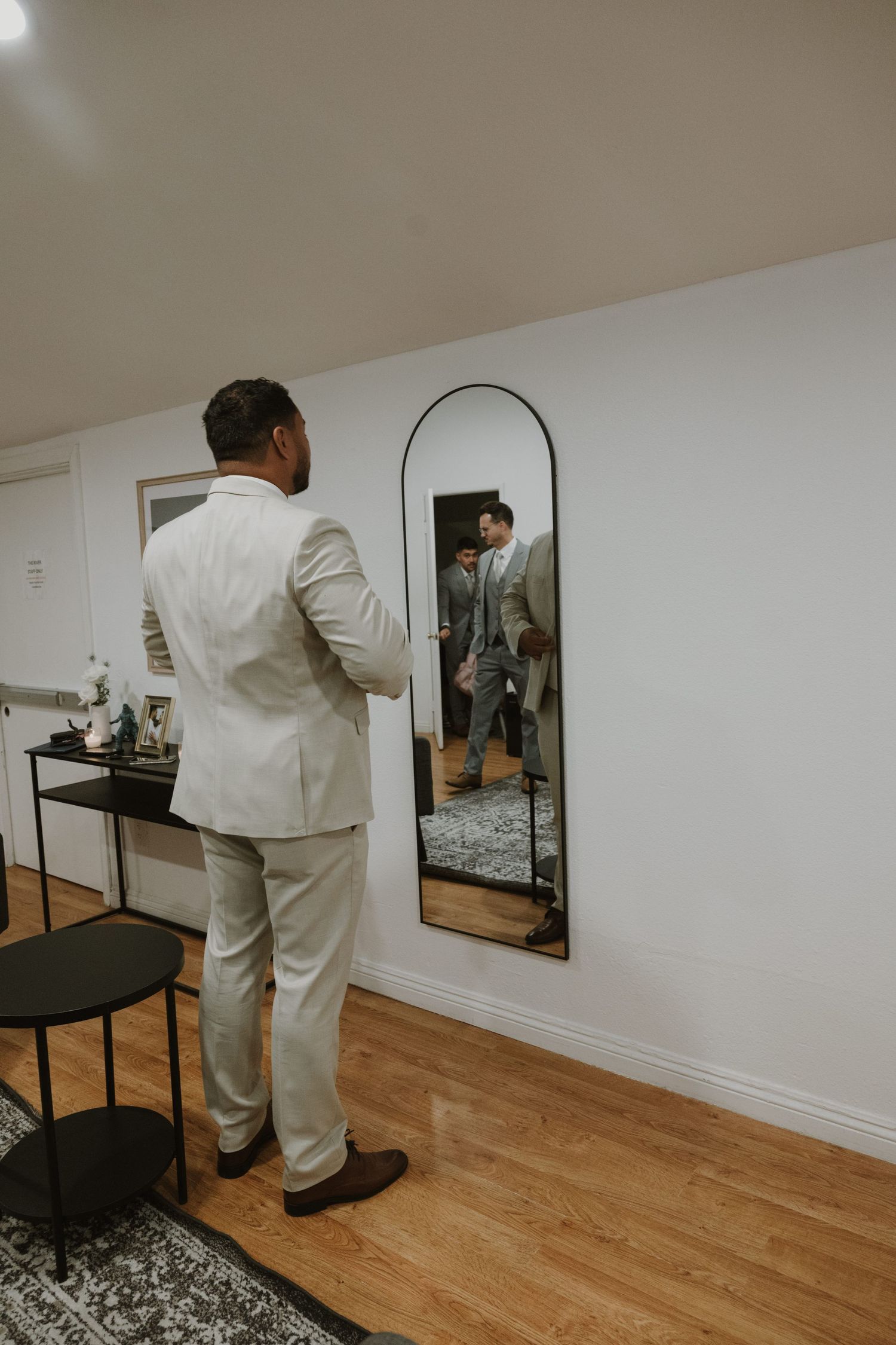 A groom in a beige suit checks his appearance in an arched floor mirror in a modern room with hardwood floors.