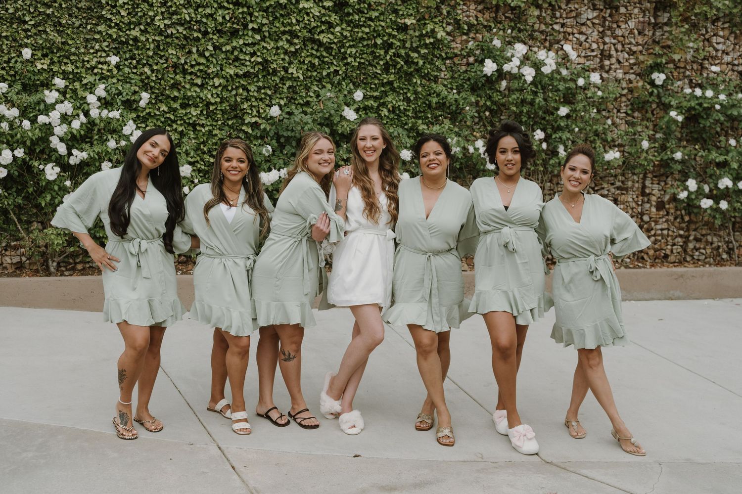 Six bridesmaids in sage green ruffle robes posing with bride in white robe before a lush green ivy wall.