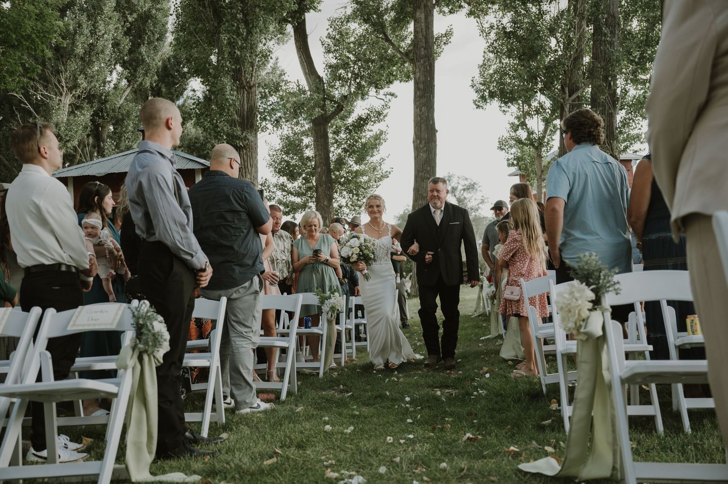 Wedding guests gather under tall trees for an outdoor ceremony with white folding chairs arranged on the grass.