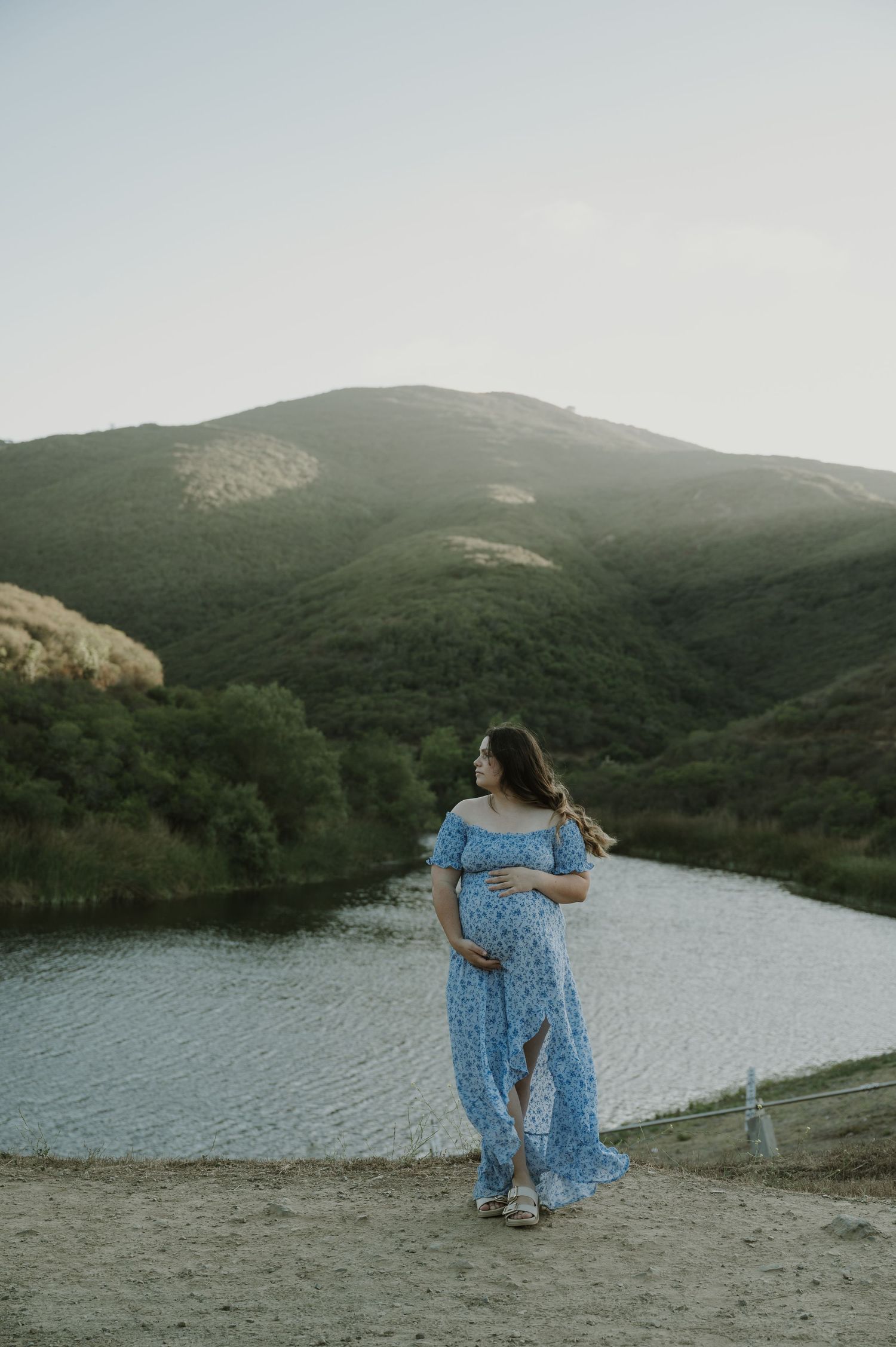 A couple stands together near a serene lake surrounded by rolling hills.