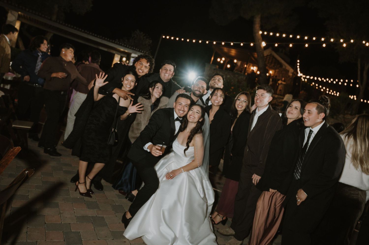 Wedding reception crowd dances and celebrates under warm string lights.