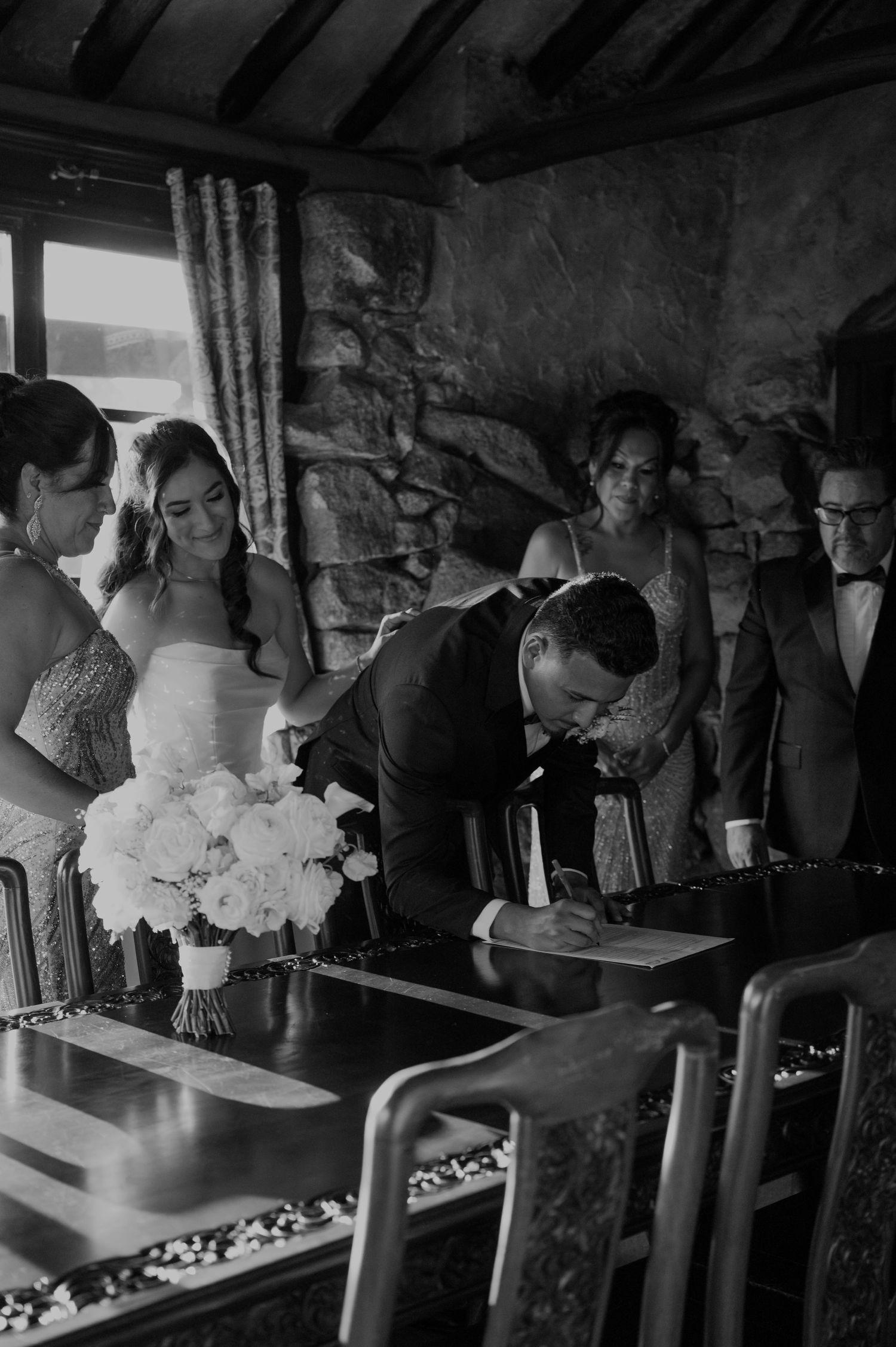 Wedding ceremony guests gathered around a table for document signing in a rustic stone room.