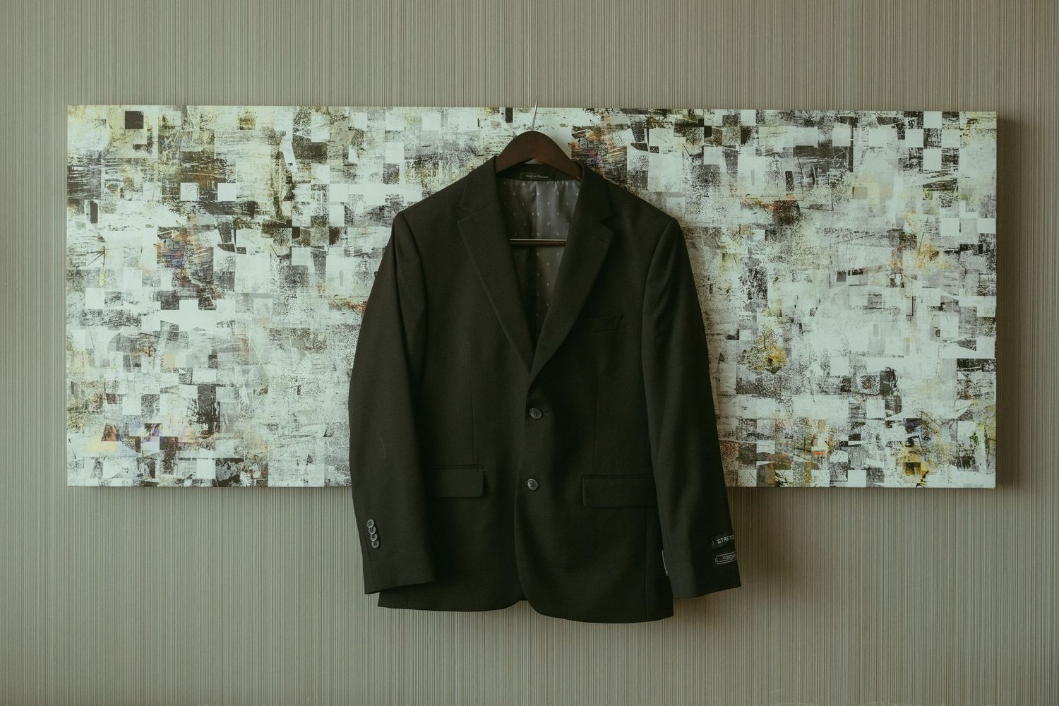 A dark brown men's suit jacket hanging on a wooden hanger against a decorative mosaic wall art backdrop.
