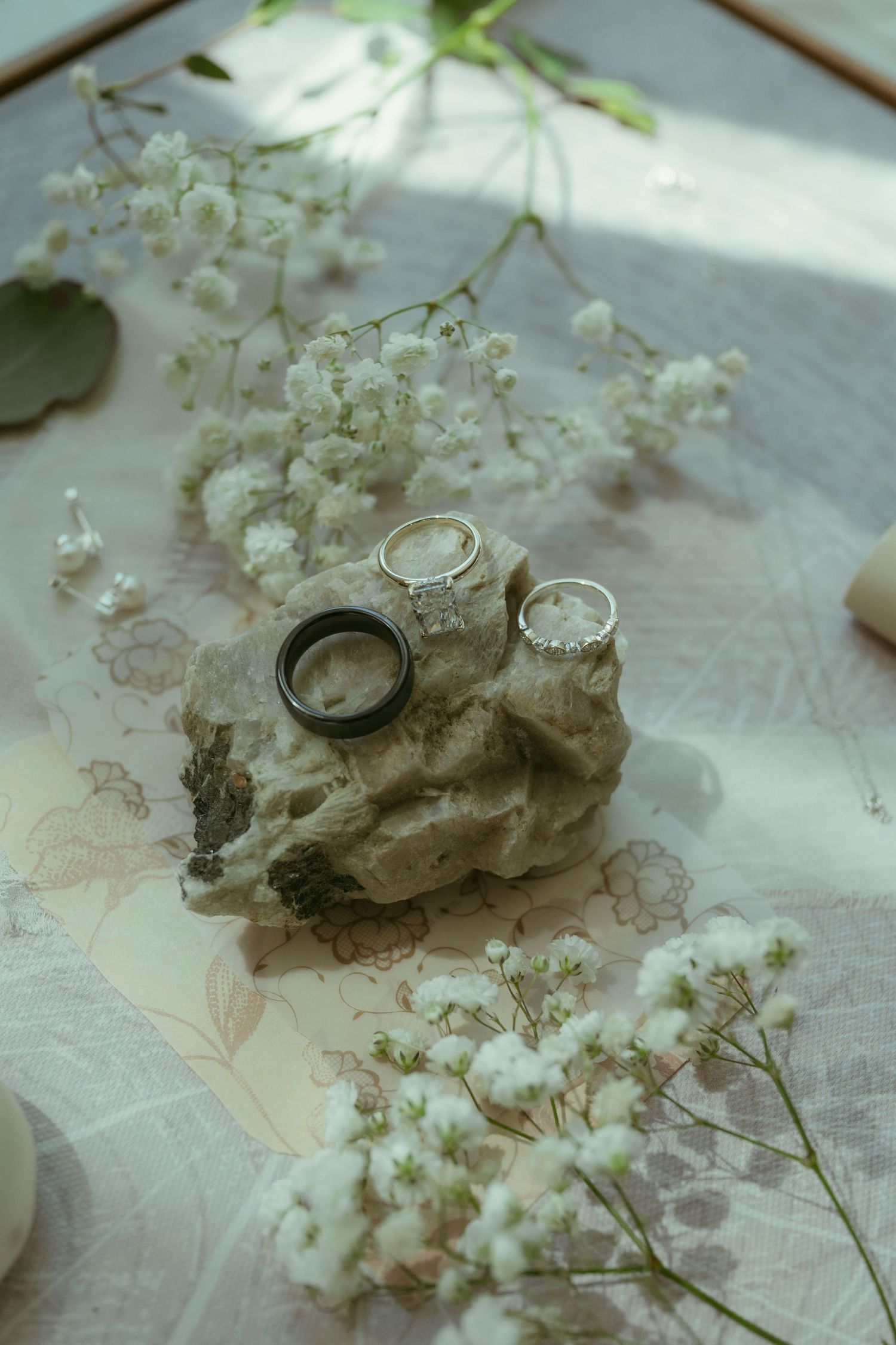 Three elegant wedding rings displayed on a rustic stone surrounded by delicate white baby's breath flowers on a floral tablecloth.