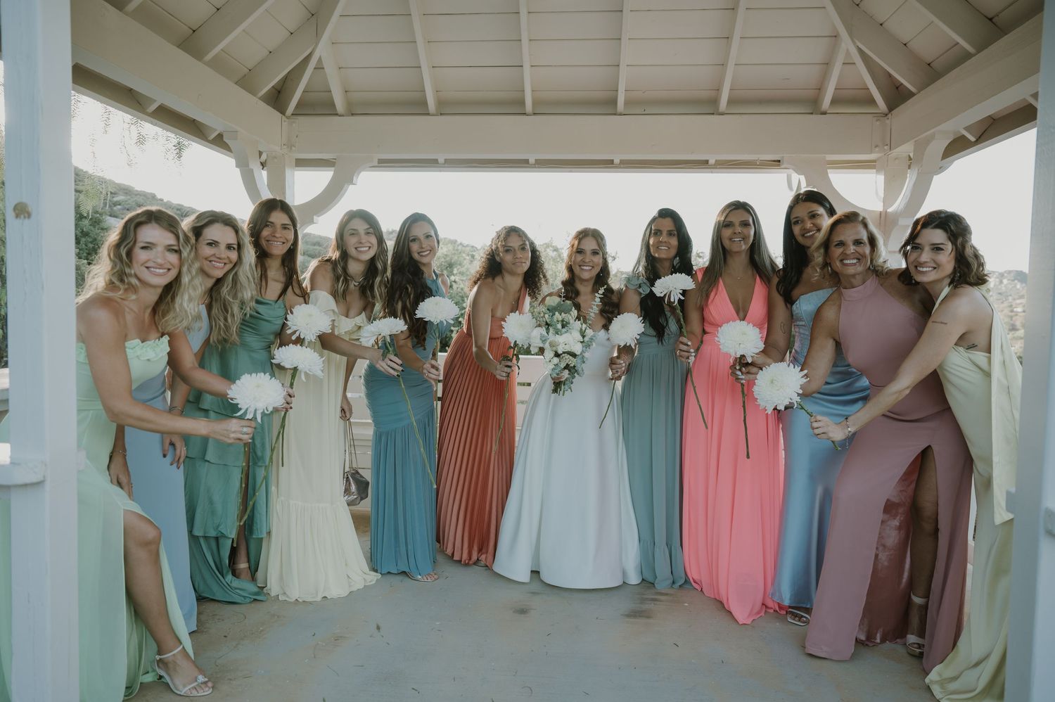 Bride poses with bridesmaids in mismatched colorful gowns holding white bouquets under white gazebo.
