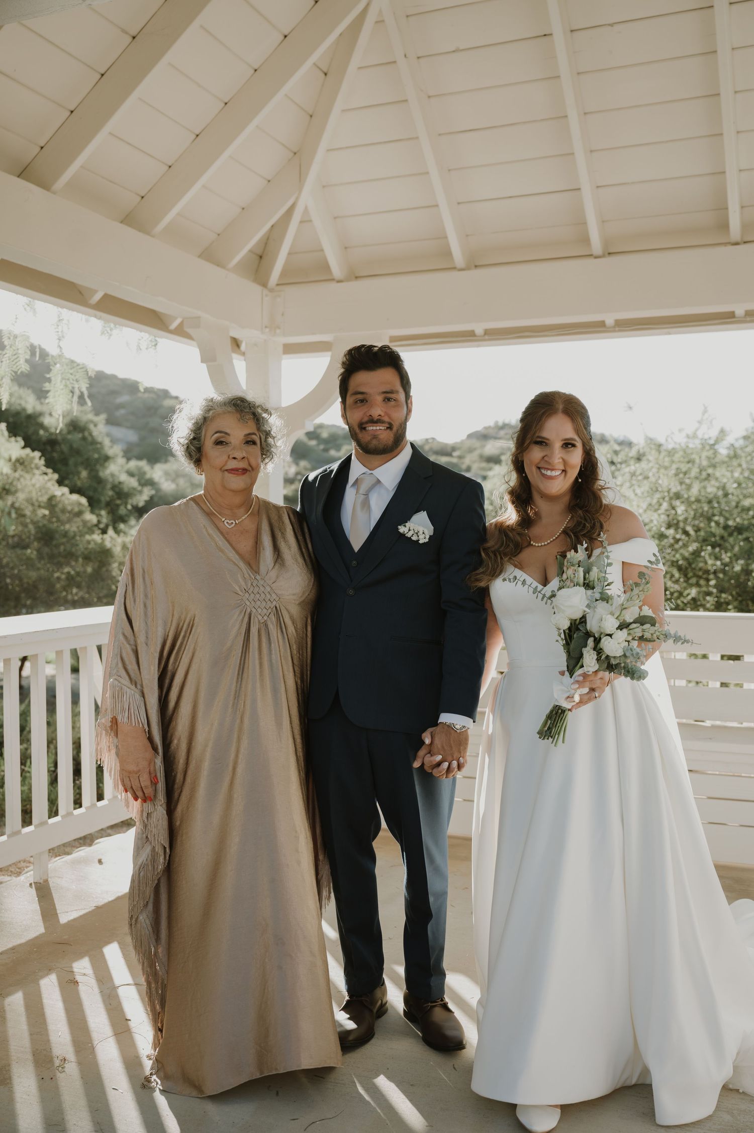 A groom in navy suit stands with his bride in white gown and an older woman in a gazebo.