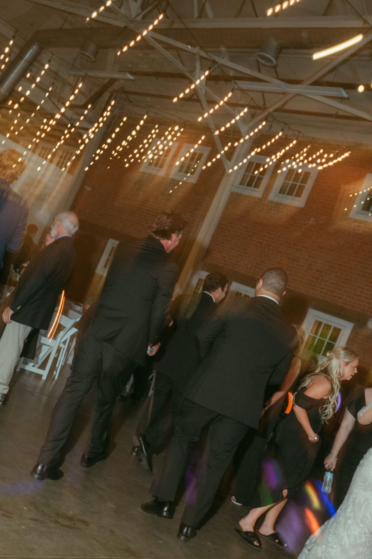 Wedding guests dancing under warm string lights in a rustic brick reception venue at night.