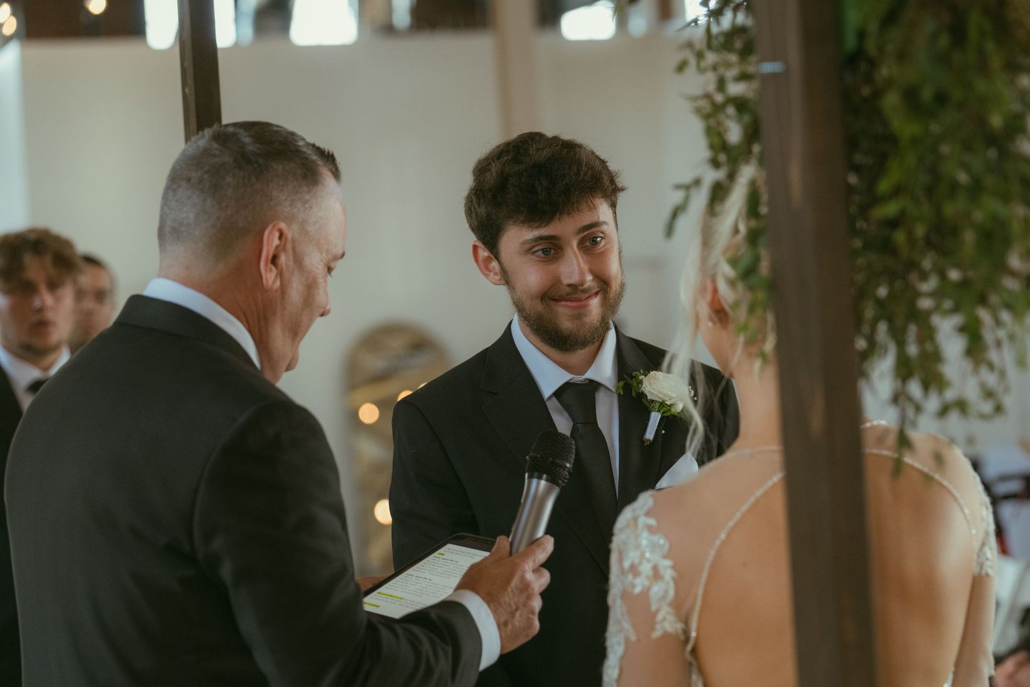 Groom smiling at bride during wedding ceremony while officiant reads vows in elegant venue.