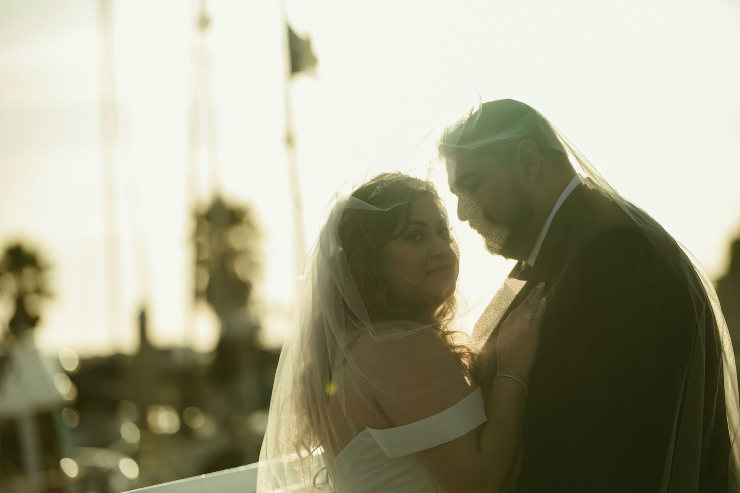 Romantic backlit wedding photo of bride and groom embracing at sunset near a marina with palm trees.