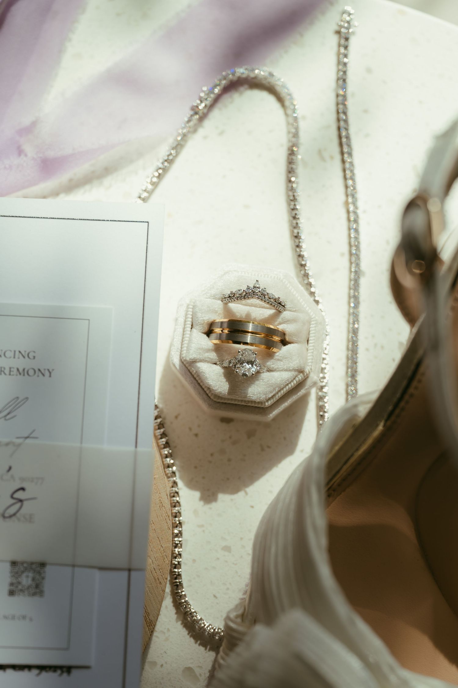 Wedding rings in a velvet box with diamond necklace, ceremony program, and bridal accessories on marble surface.