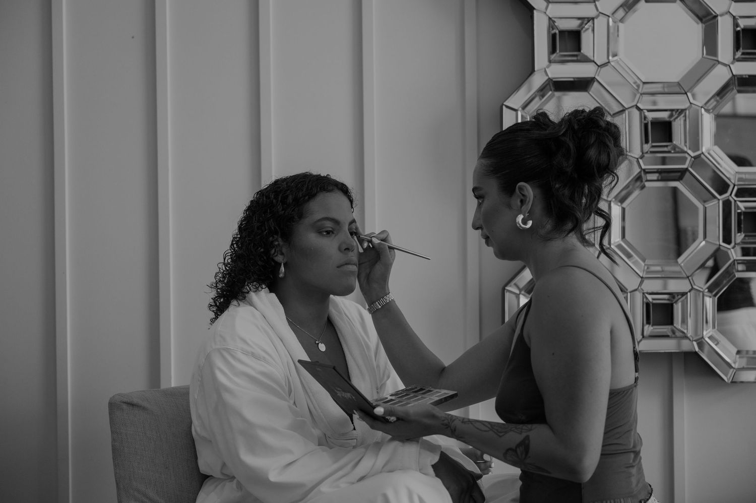 A makeup artist applies makeup to a seated woman in a white robe in a black and white photo.