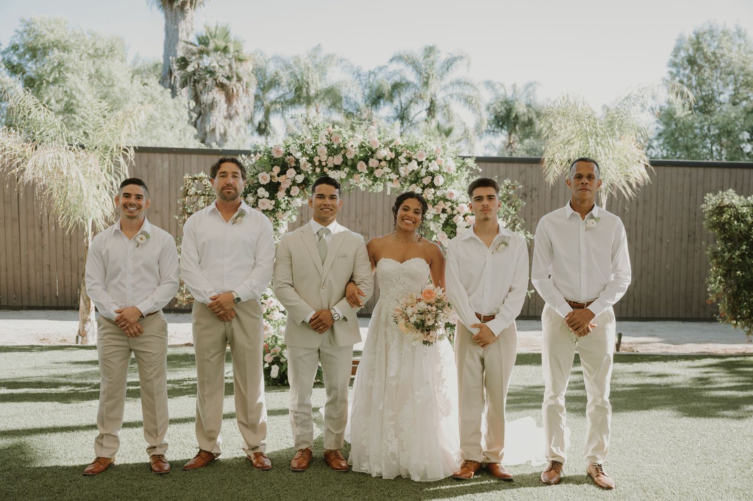 Bride in white gown poses with four groomsmen in white shirts and khaki pants before floral arch.