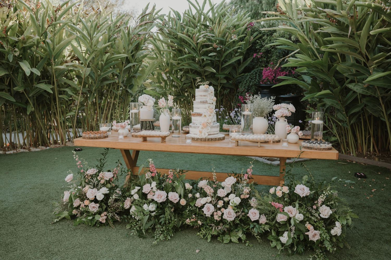 Rustic wooden wedding dessert table with tiered cake and pink floral arrangements on green lawn.