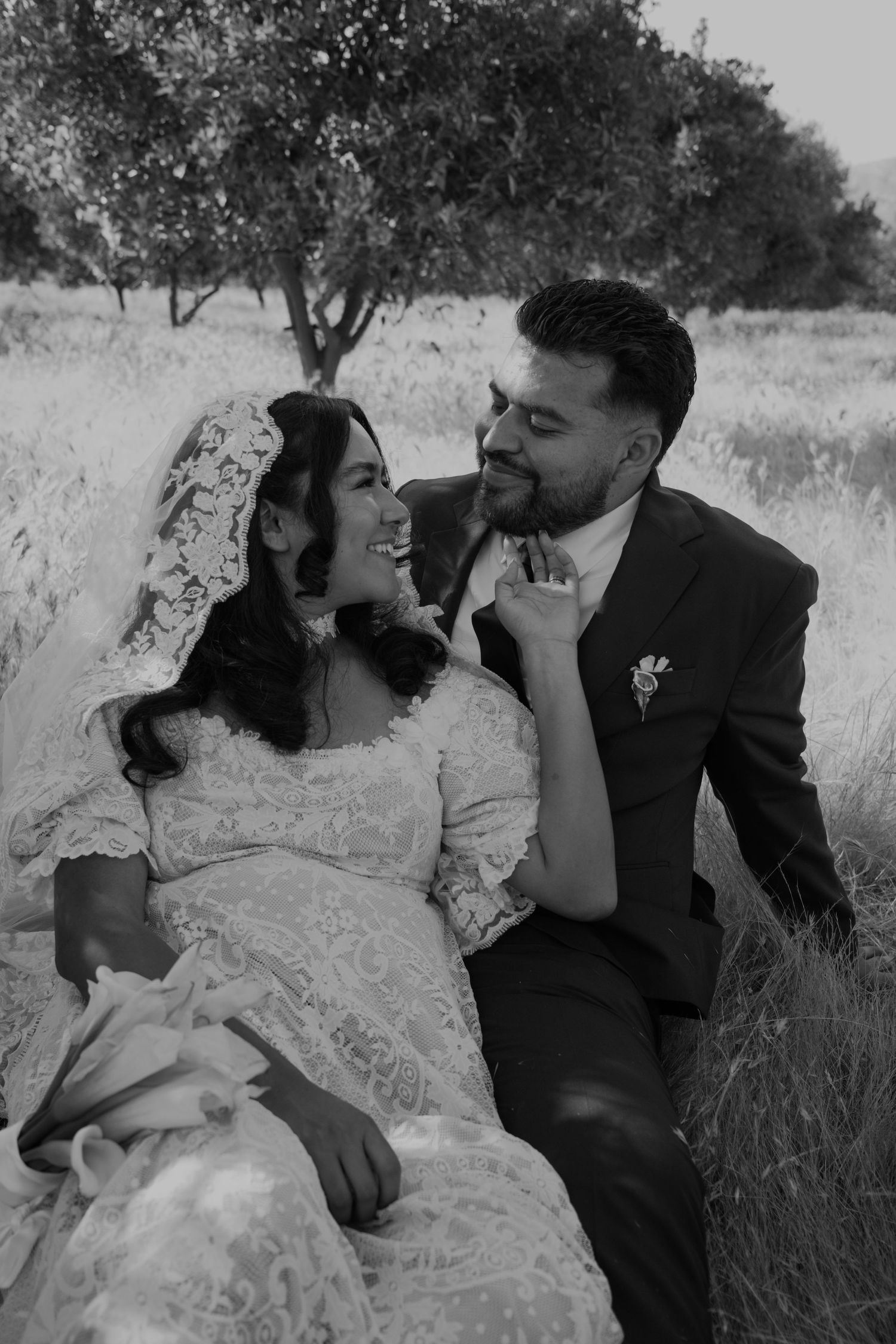 Black and white photo of a smiling bride and groom sitting together in a field, sharing a joyful moment on their wedding day.