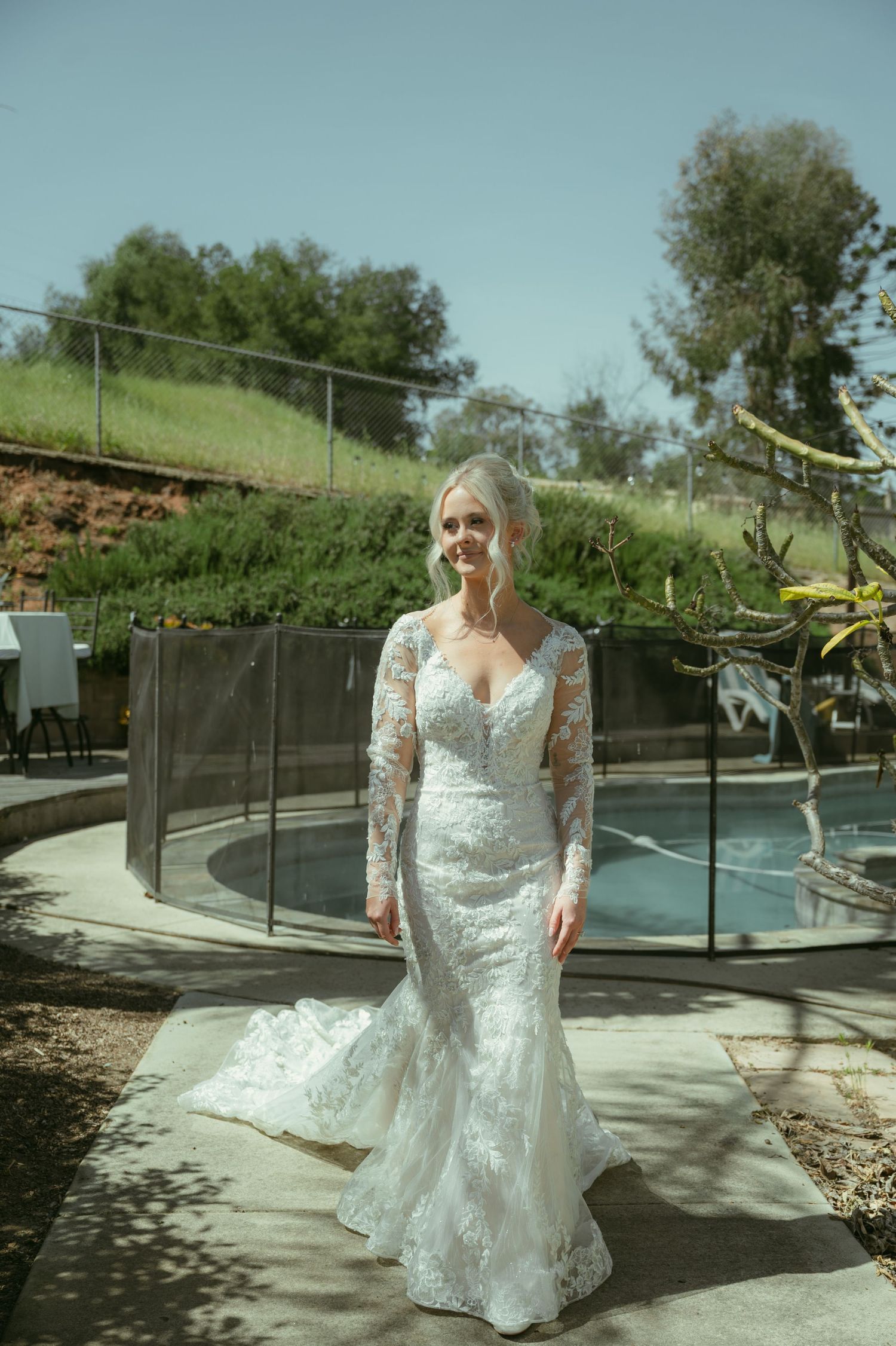 A bride in a long-sleeve lace mermaid wedding gown poses outdoors near a pool on a sunny day.