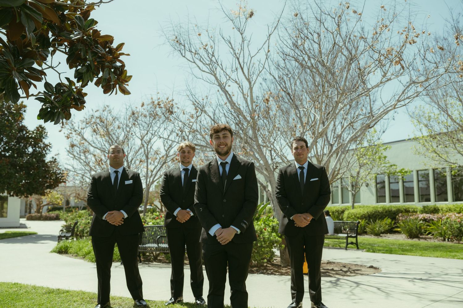 Groom and three groomsmen in matching black suits standing outdoors on wedding day.
