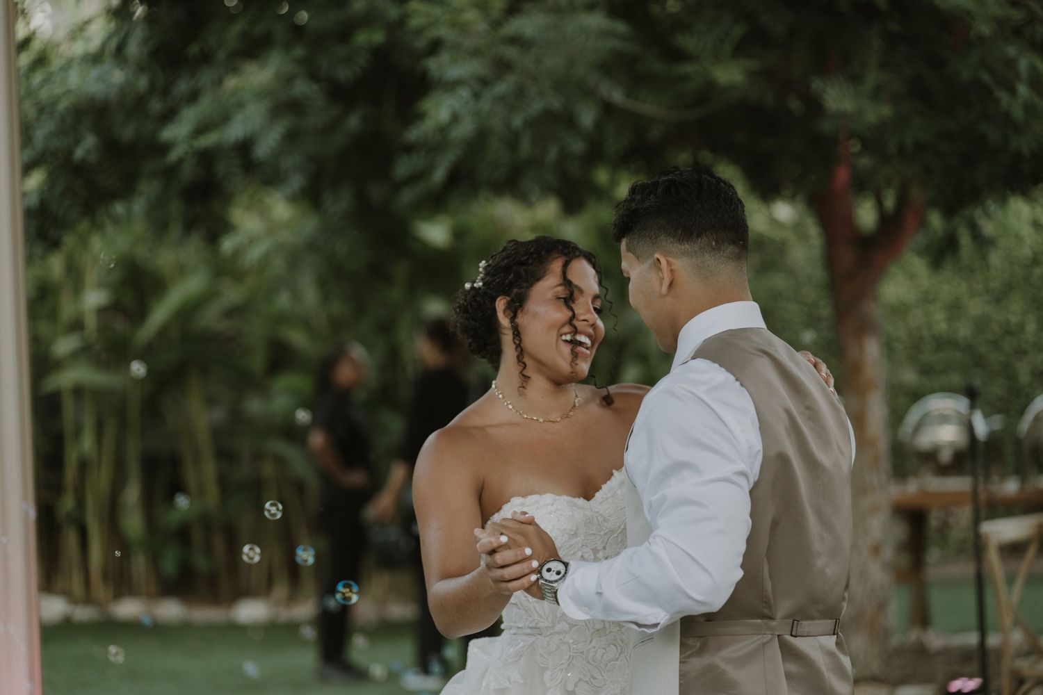 Bride and groom share first dance outdoors surrounded by bubbles and lush green trees at wedding reception.