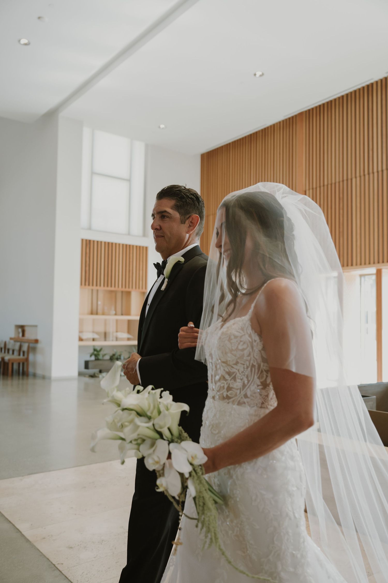 Bride in lace gown and veil holds white bouquet beside escort in black tuxedo at church wedding.