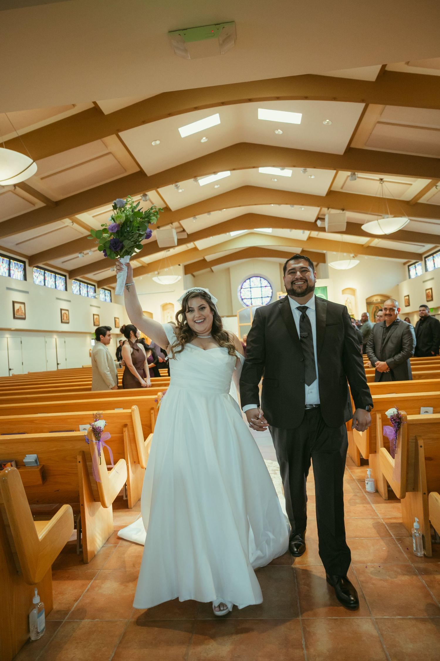 Joyful newlyweds walk down church aisle after wedding ceremony, bride raising bouquet in celebration.