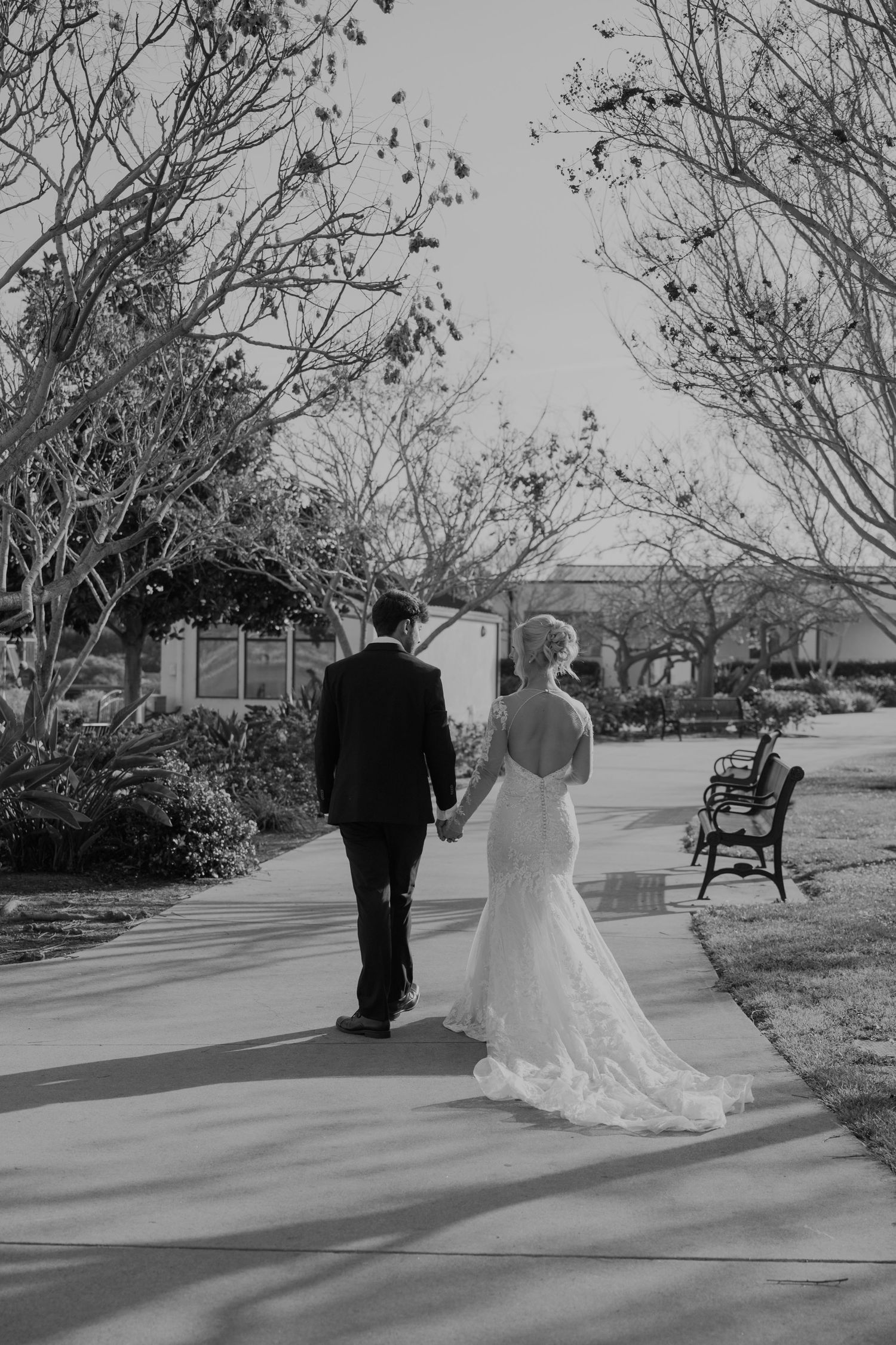 Black and white photo of bride and groom walking hand in hand down a tree-lined path on their wedding day.