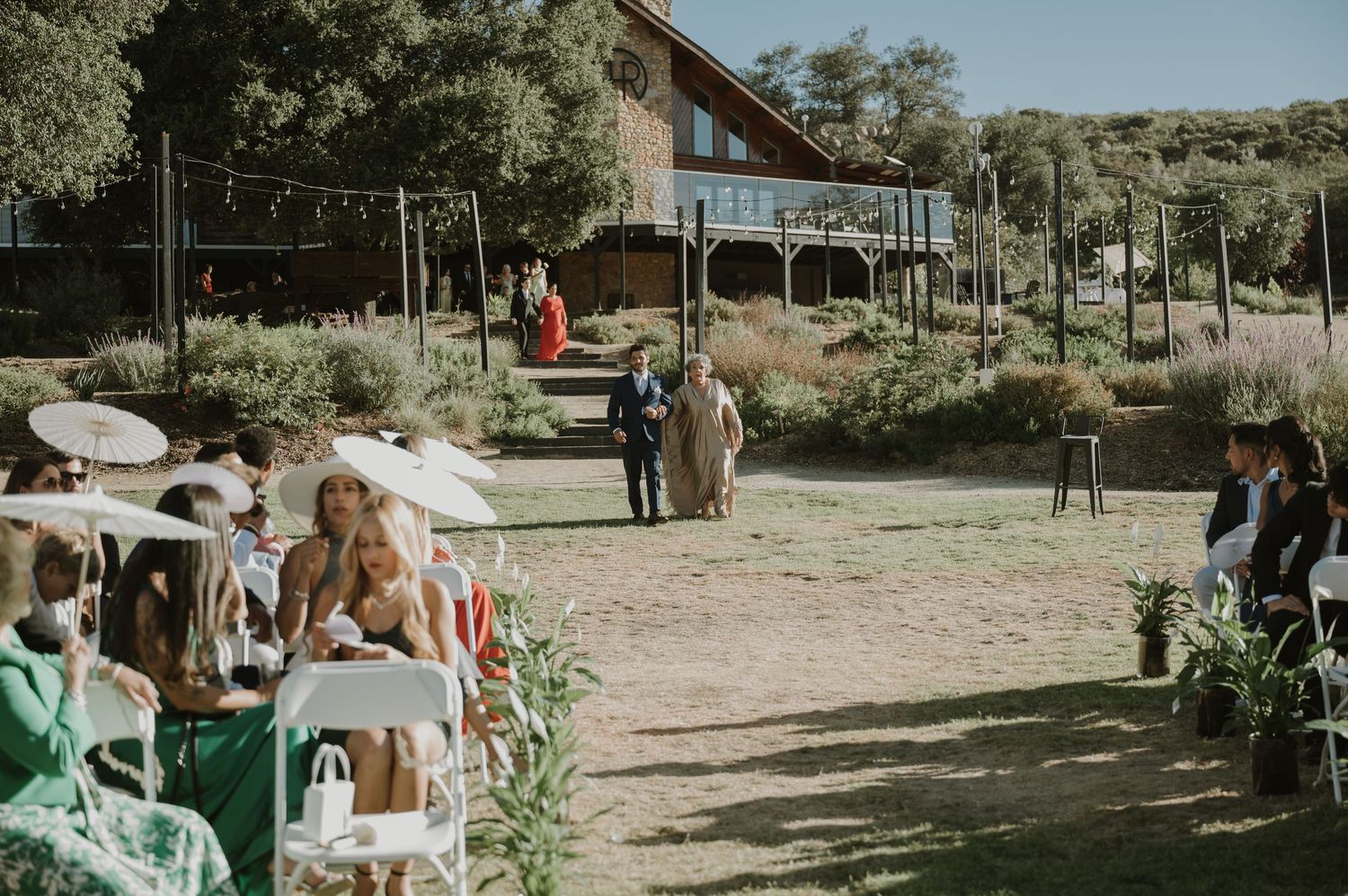 Outdoor wedding ceremony with guests seated on chairs in a garden setting with a rustic wooden venue in the background.