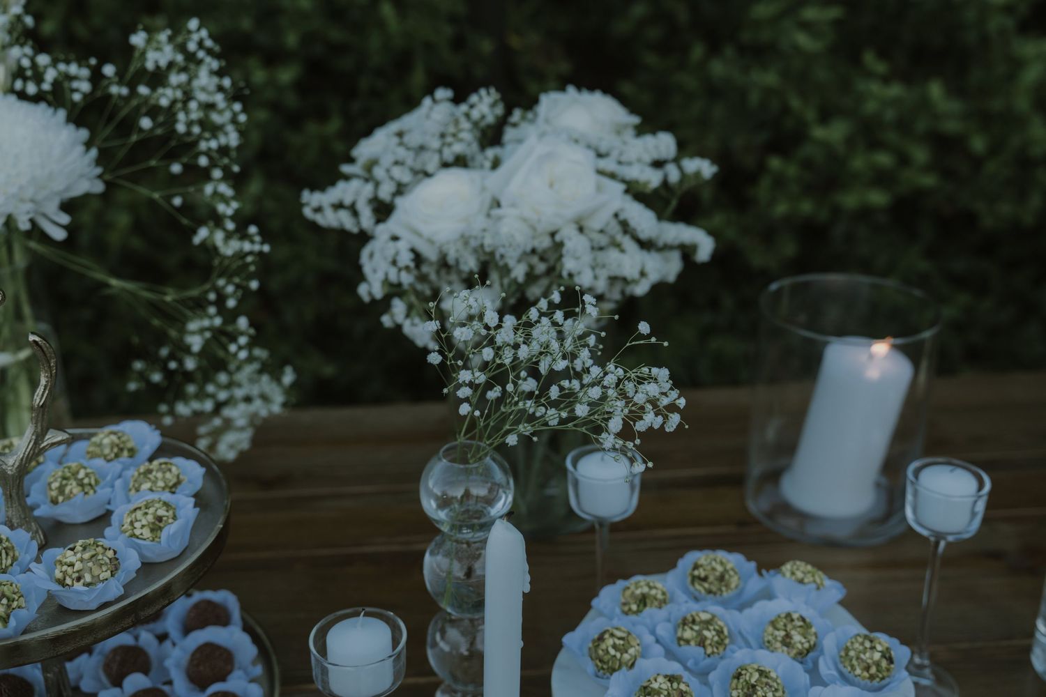 Elegant outdoor dessert table with white floral arrangements, candles, and decorated treats on a rustic wooden surface.