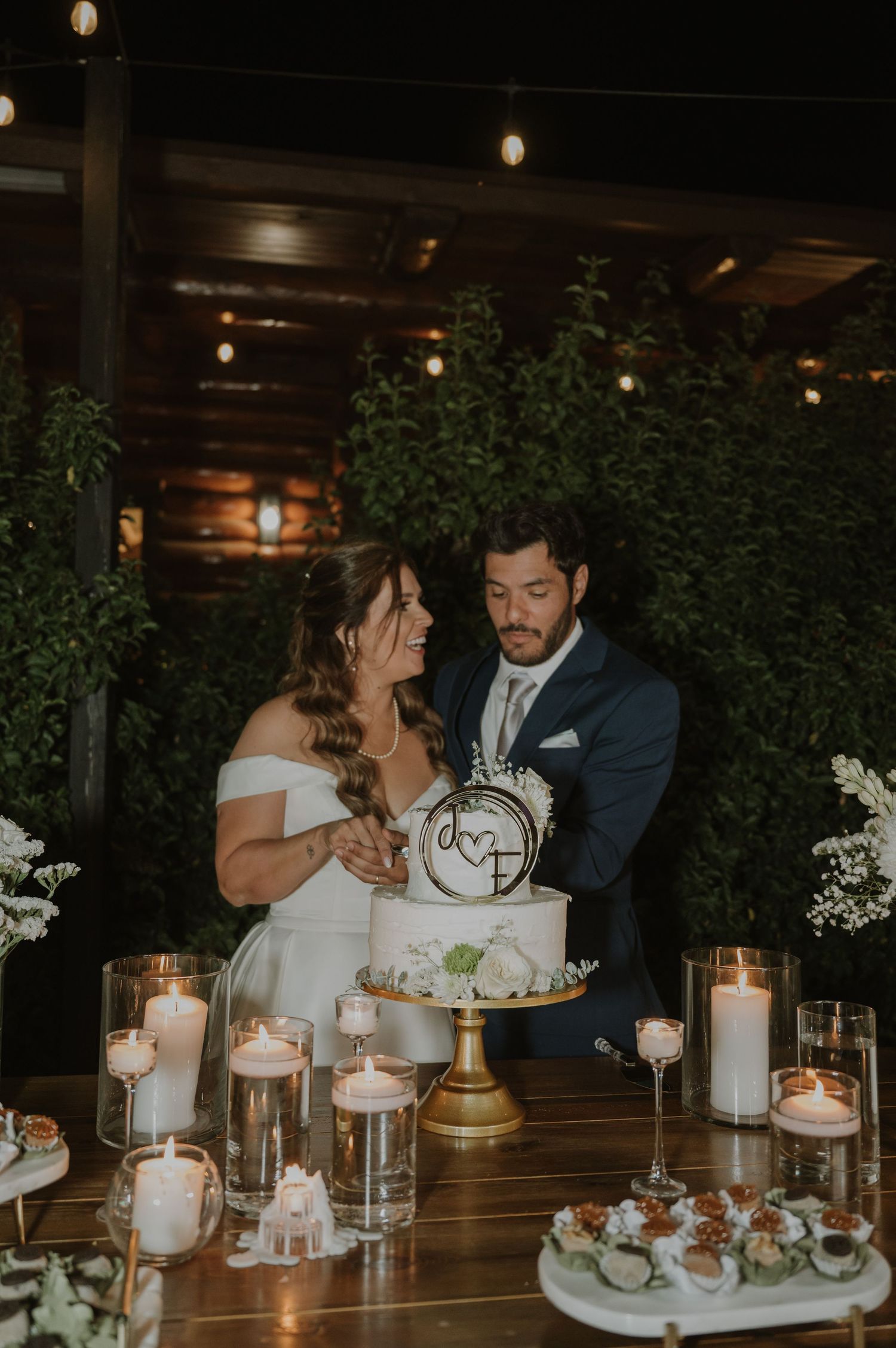 Bride and groom cutting wedding cake outdoors at night surrounded by candles and string lights.