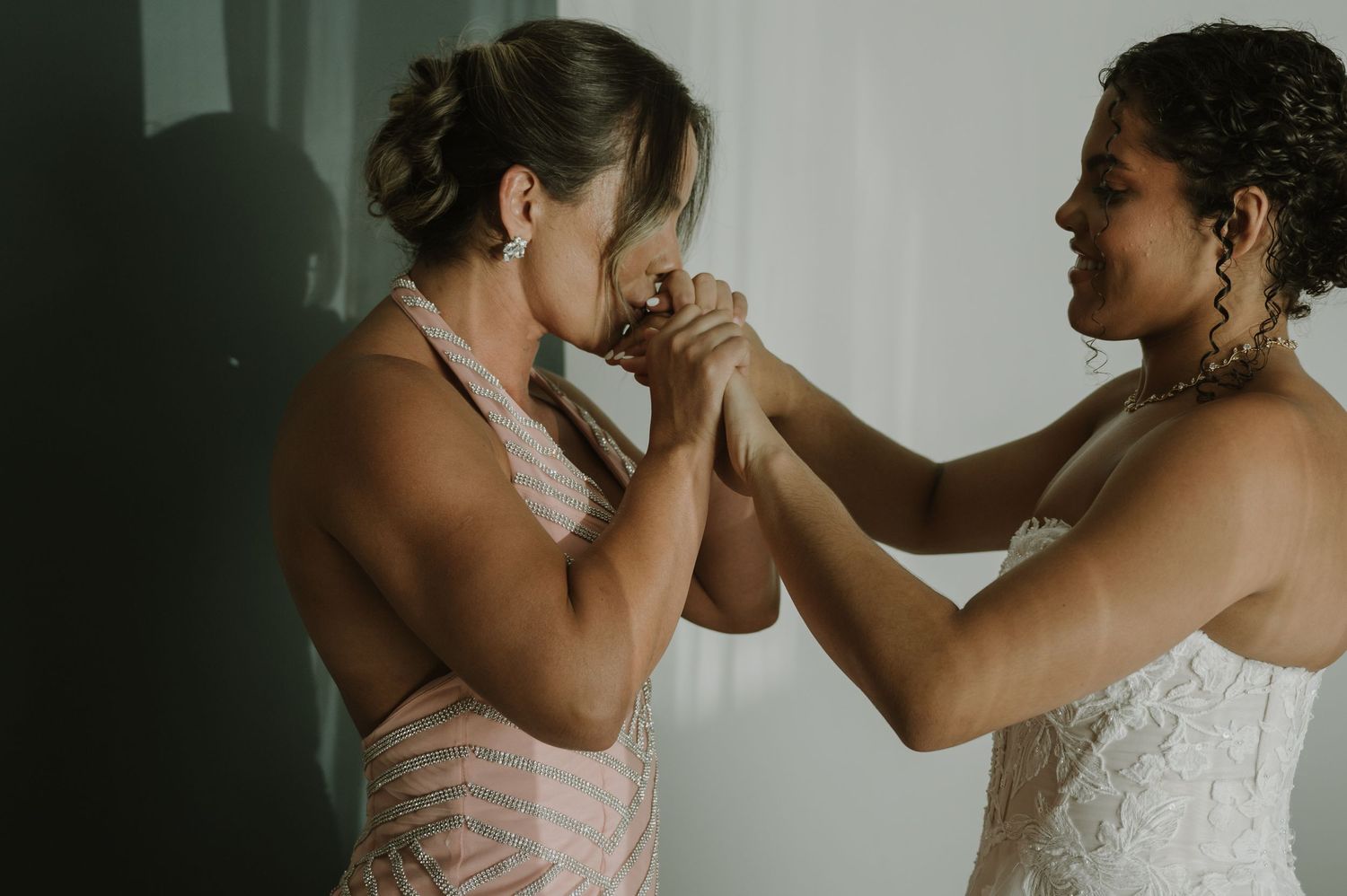 Emotional bride in white lace gown holds hands with woman in pink dress during wedding preparations.