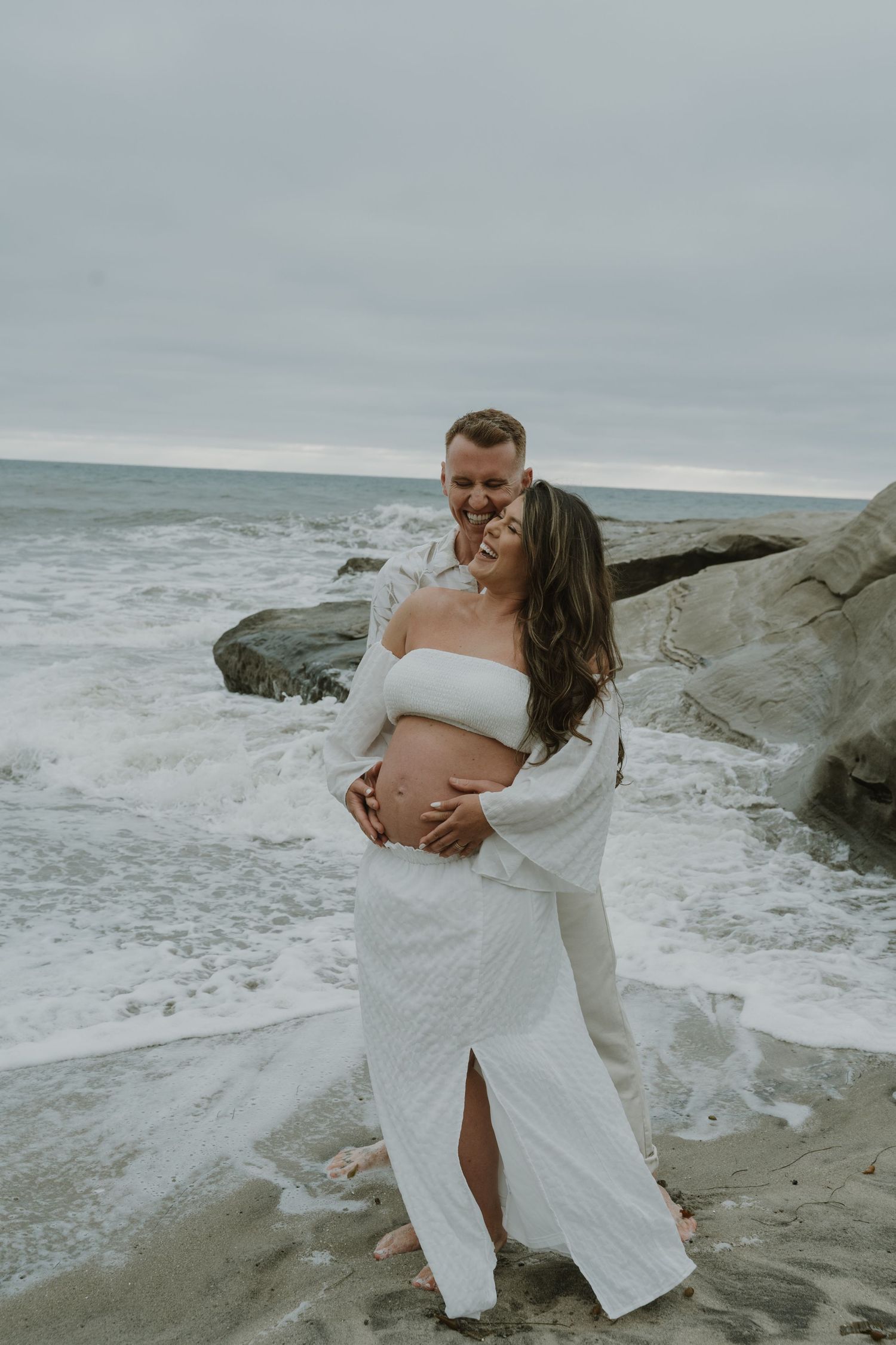 Romantic maternity photography session along a rocky beach with crashing waves.