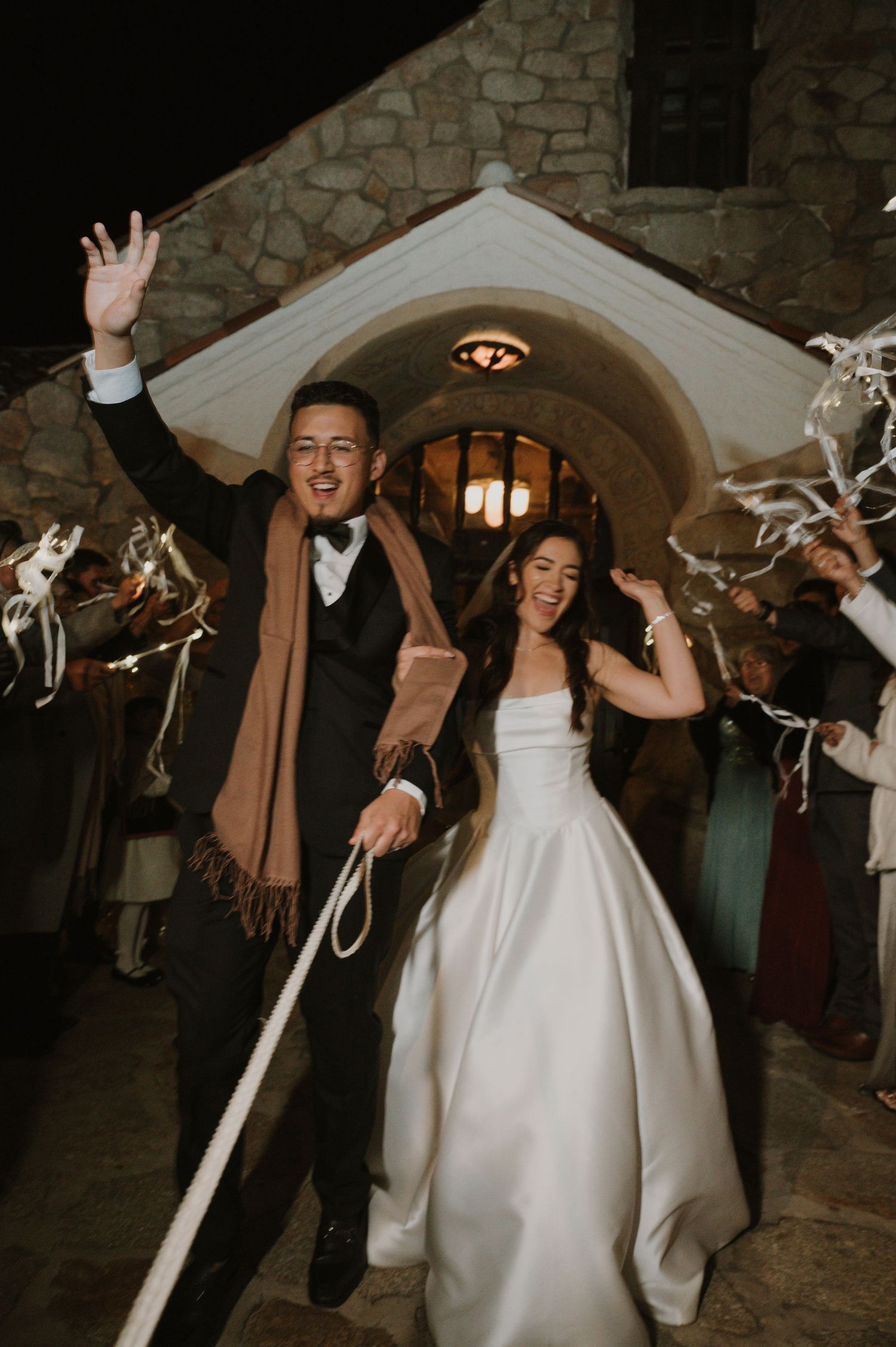 Joyful wedding reception entrance with guests waving ribbons under stone archways.