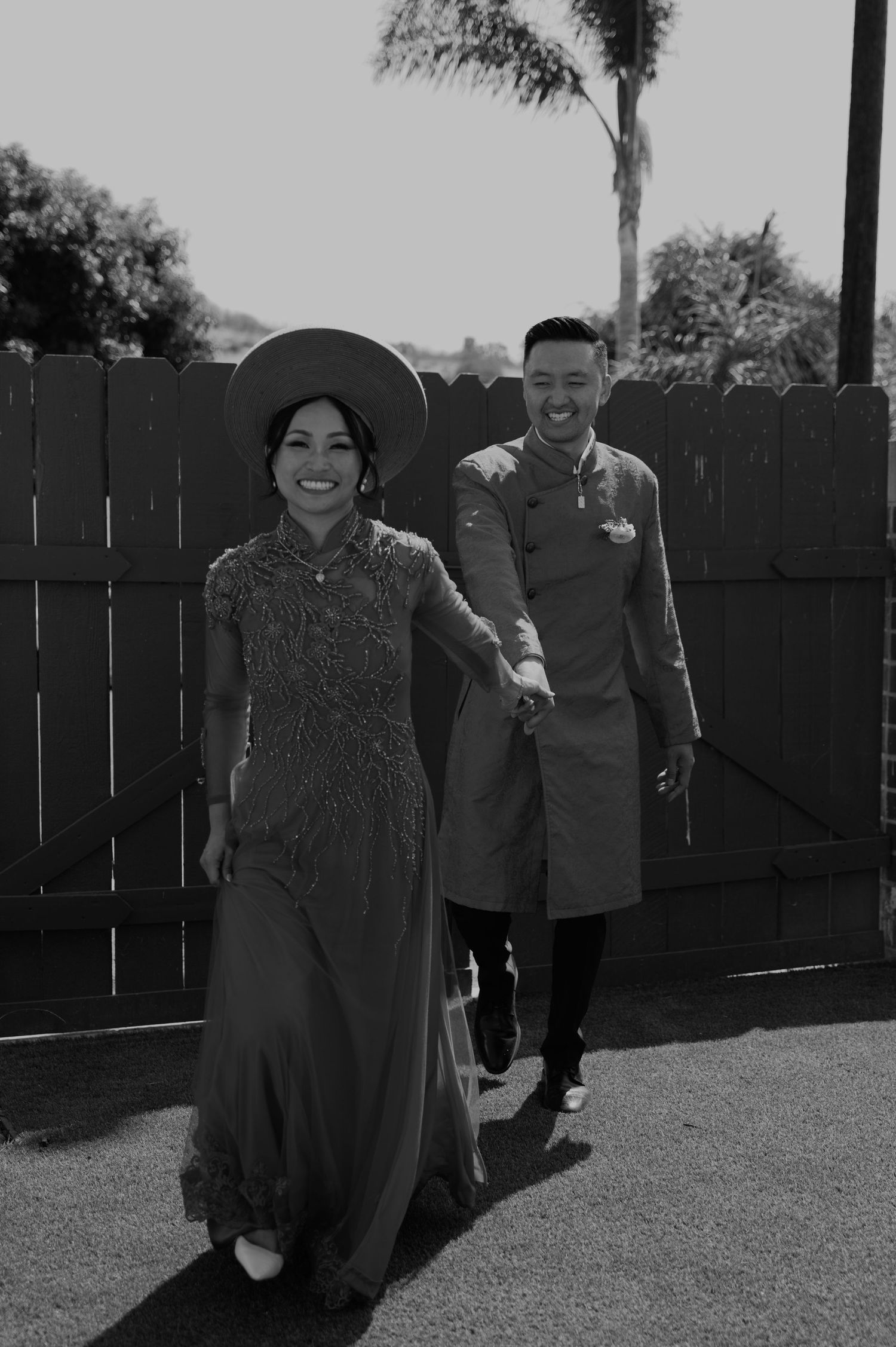 Black and white photo of couple in traditional formal attire posing by wooden fence.