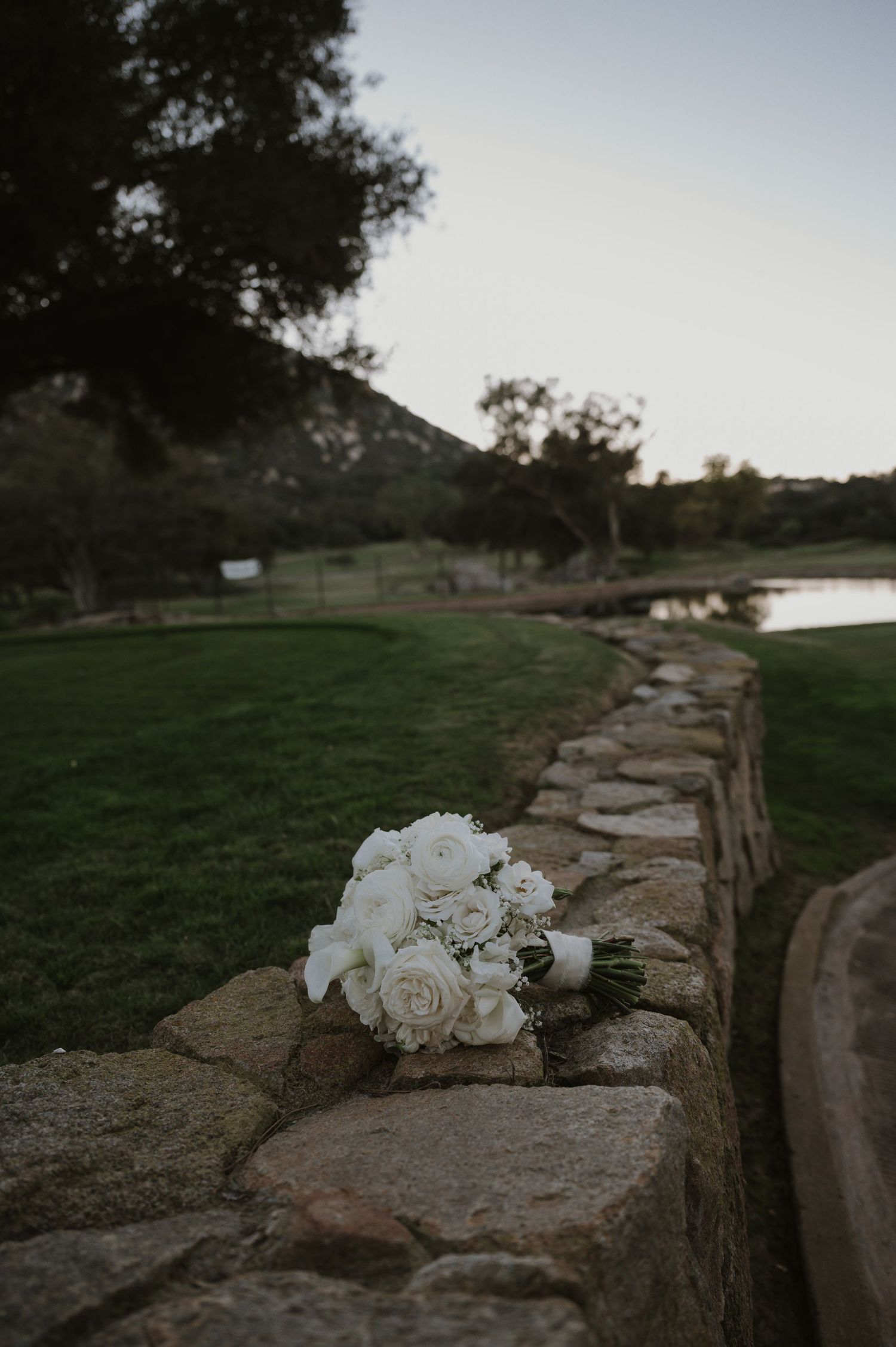 A white wedding bouquet rests on a rustic stone wall with a scenic mountain landscape and pond in the background.