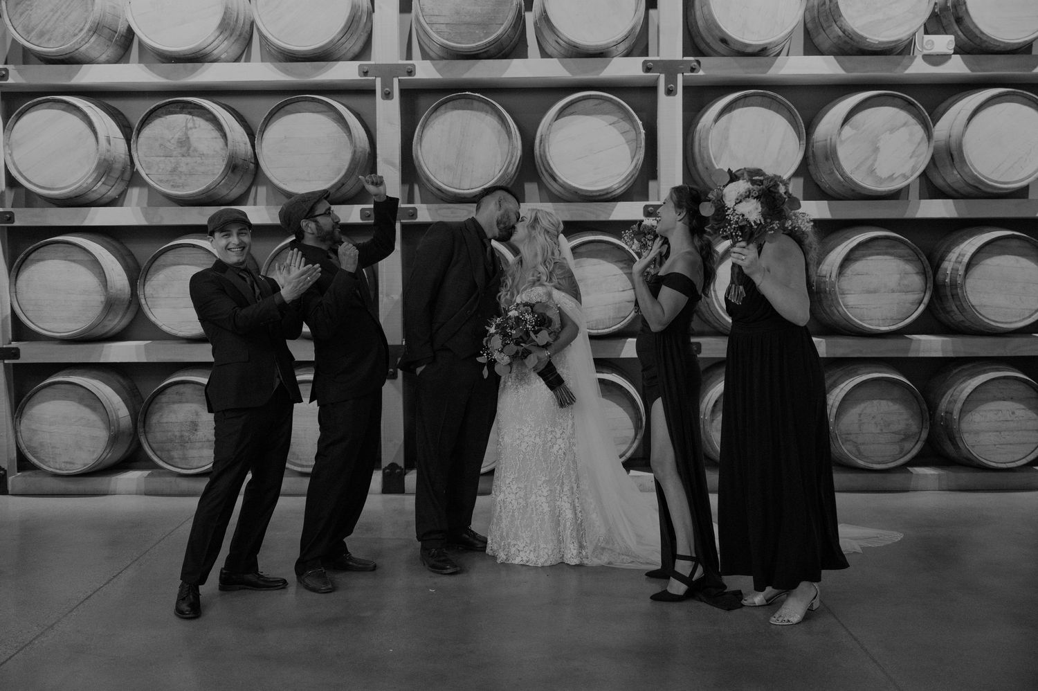 Wedding group photo in front of stacked wine barrels in a rustic cellar.