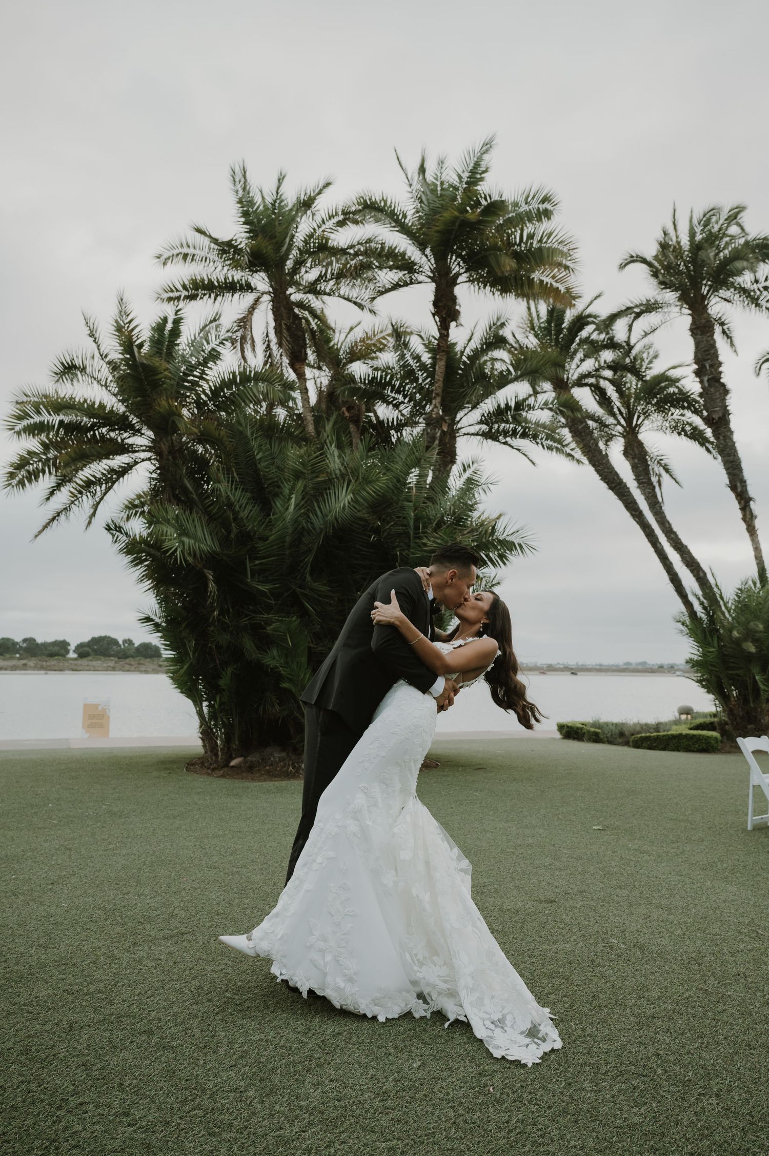 Wedding couple dances beneath palm trees on a manicured lawn overlooking the water.