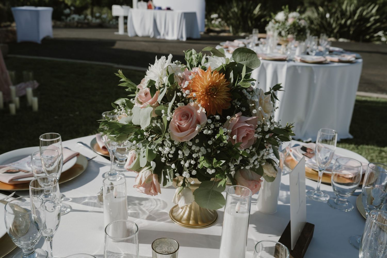 Elegant outdoor wedding reception table with gold vase centerpiece featuring pink roses, protea, and greenery.