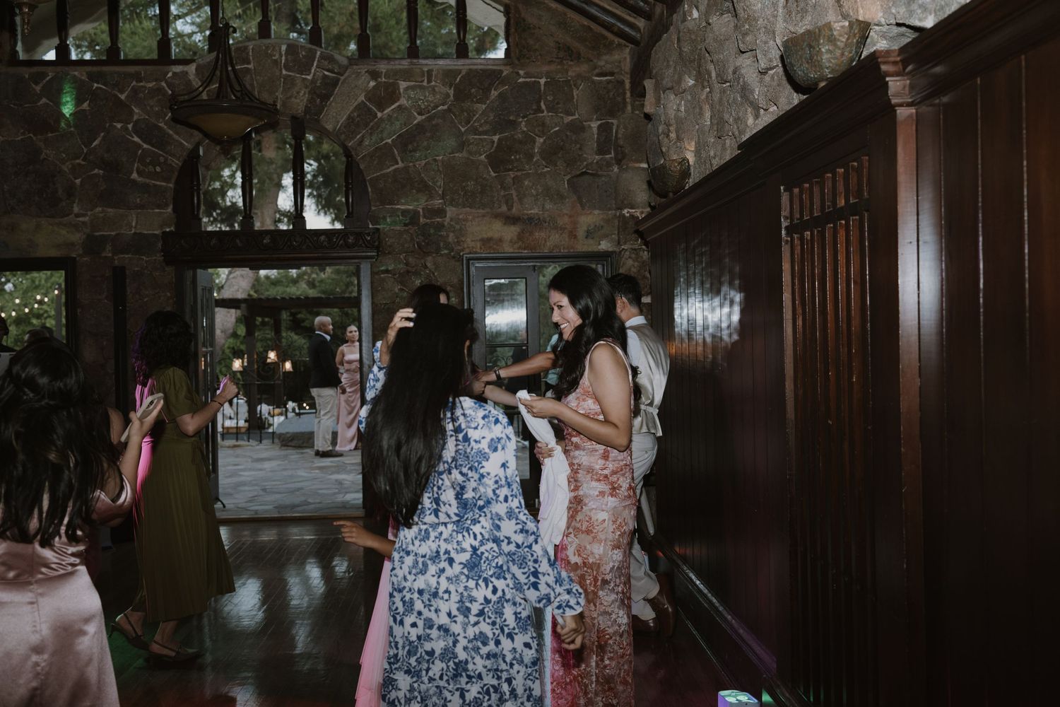 Wedding guests gather in a rustic stone venue as bridesmaids assist with dress adjustments.